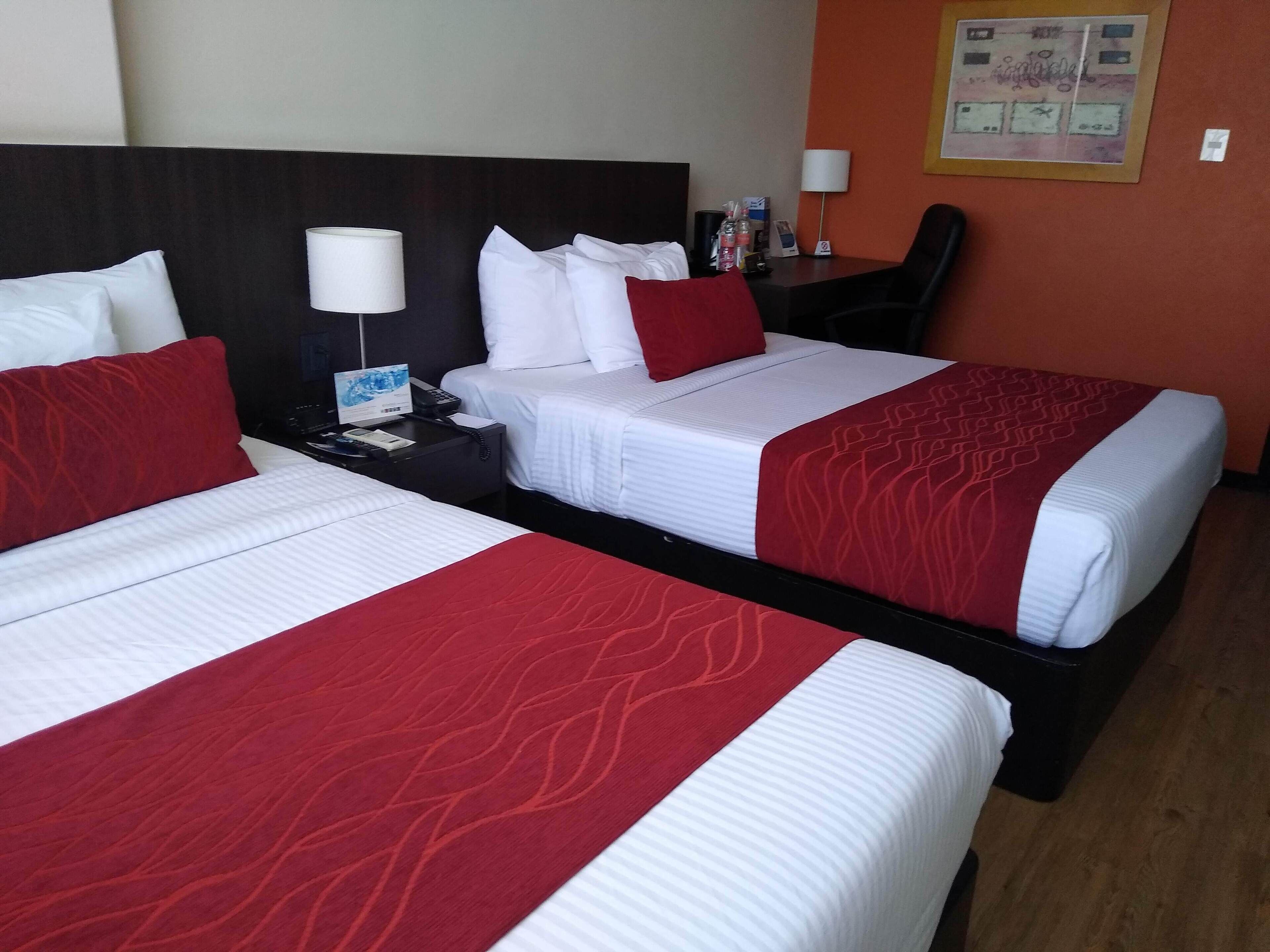Two double beds with red and white bedding, a nightstand with a lamp, a desk with a phone, and a chair.