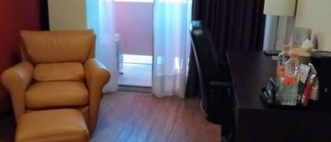 A hotel room with a wooden floor, a leather armchair, a desk with a lamp, and a view through a window with curtains.