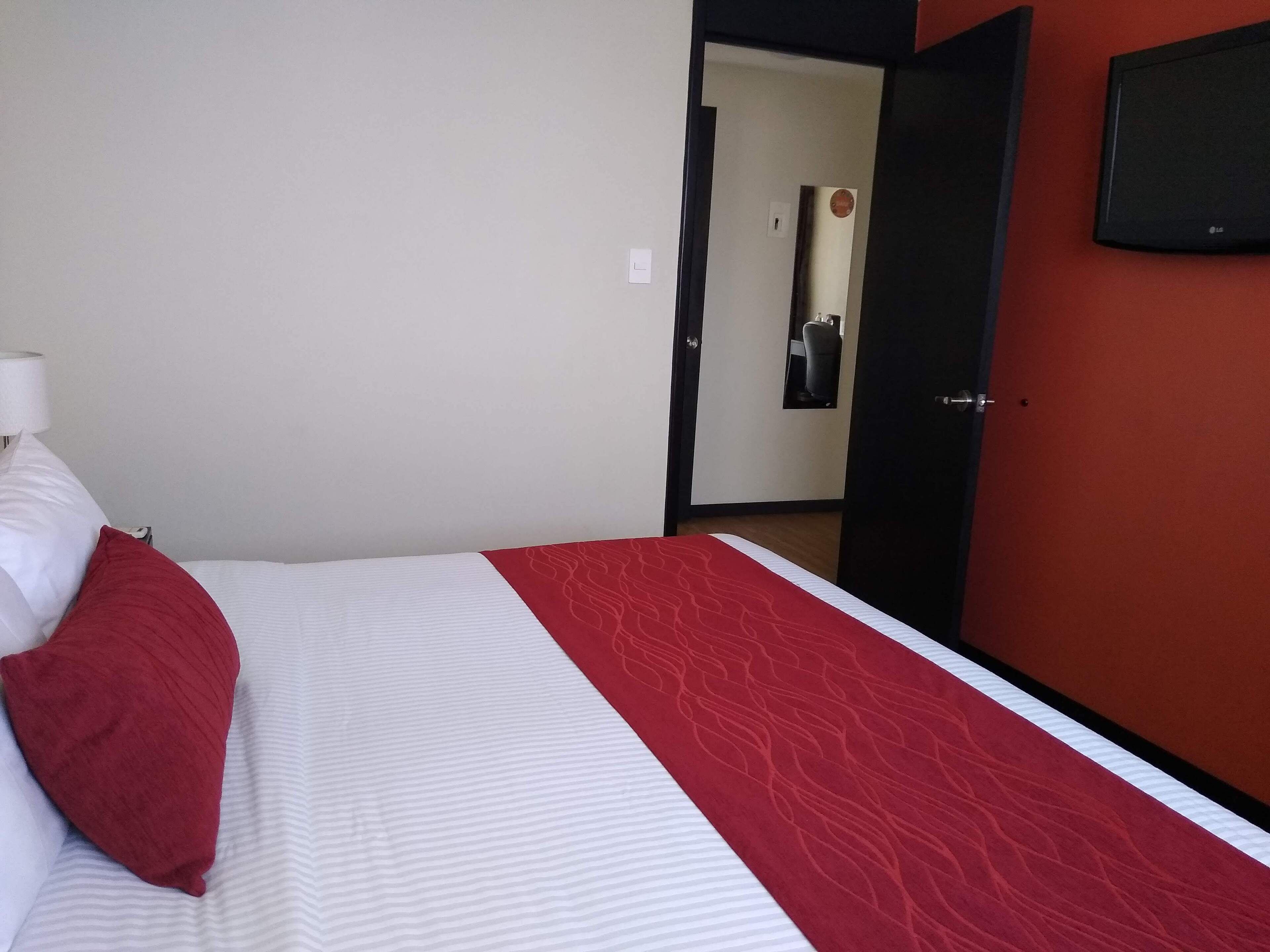 Quarto de hotel com cama, colcha listrada em vermelho e branco, travesseiro vermelho, luminária branca e TV fixada na parede.