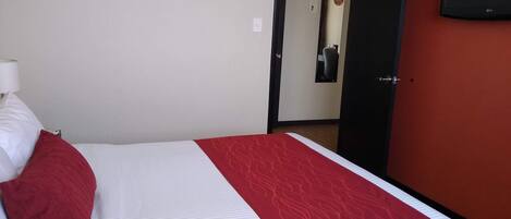 Quarto de hotel com cama, colcha listrada em vermelho e branco, travesseiro vermelho, luminária branca e TV fixada na parede.
