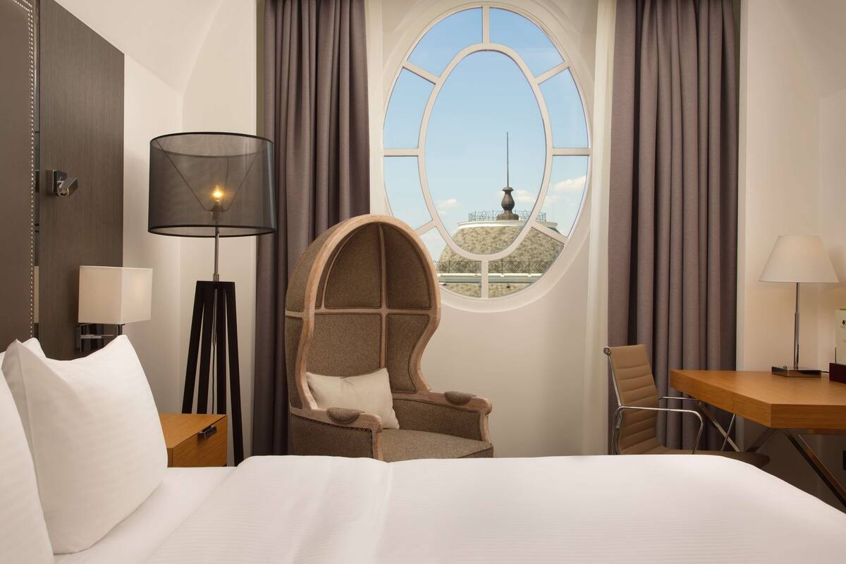Habitación de hotel con una cama grande, un ventanal redondo con vistas, una silla y un escritorio con lámpara.