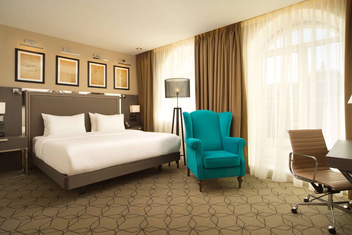 Una habitación de hotel con una cama grande, un sillón verde azulado, una silla de escritorio marrón, un escritorio y una lámpara de pie.
