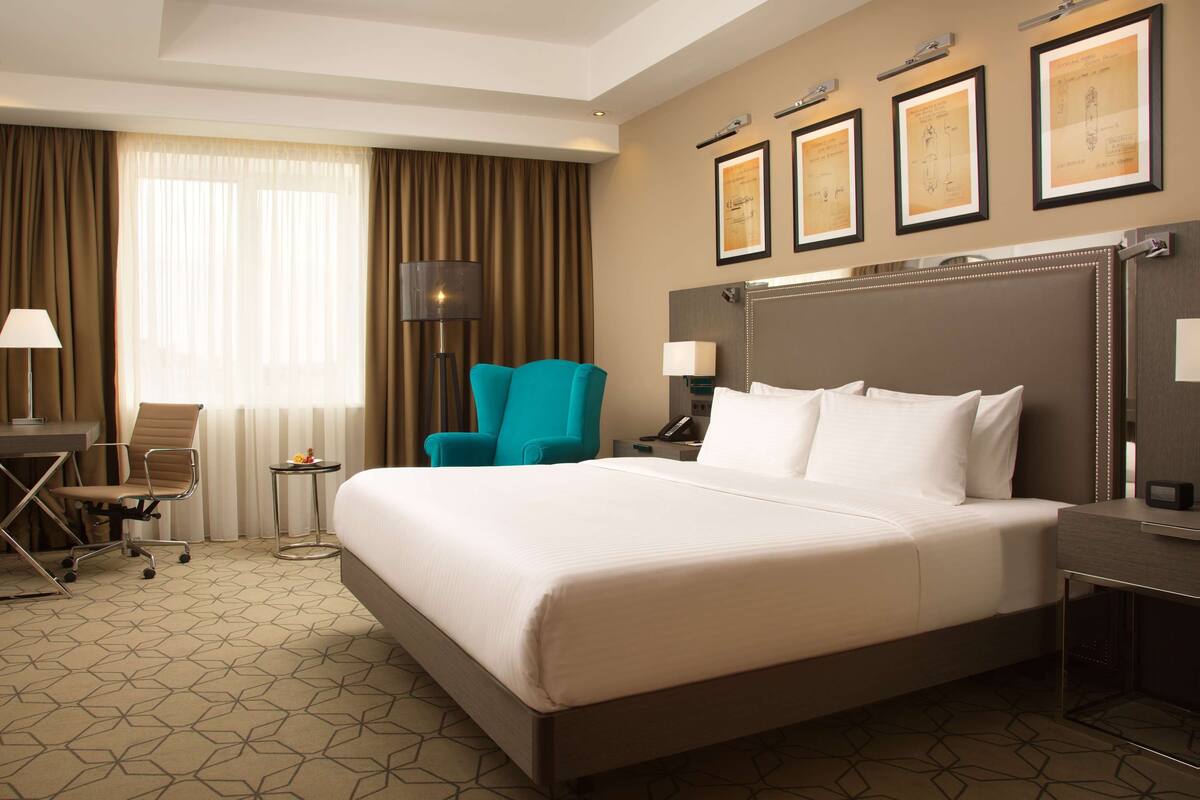 Una habitación de hotel con una cama grande, un escritorio con silla, una lámpara de pie y un sillón azul turquesa.
