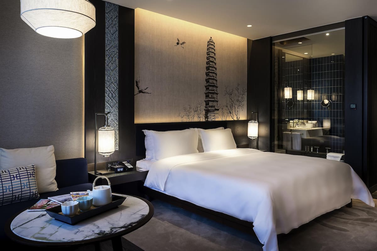Una habitación de hotel moderna con una cama grande, lámparas de noche, una mesita de noche, una mesa redonda con una bandeja y un adorno en la pared.