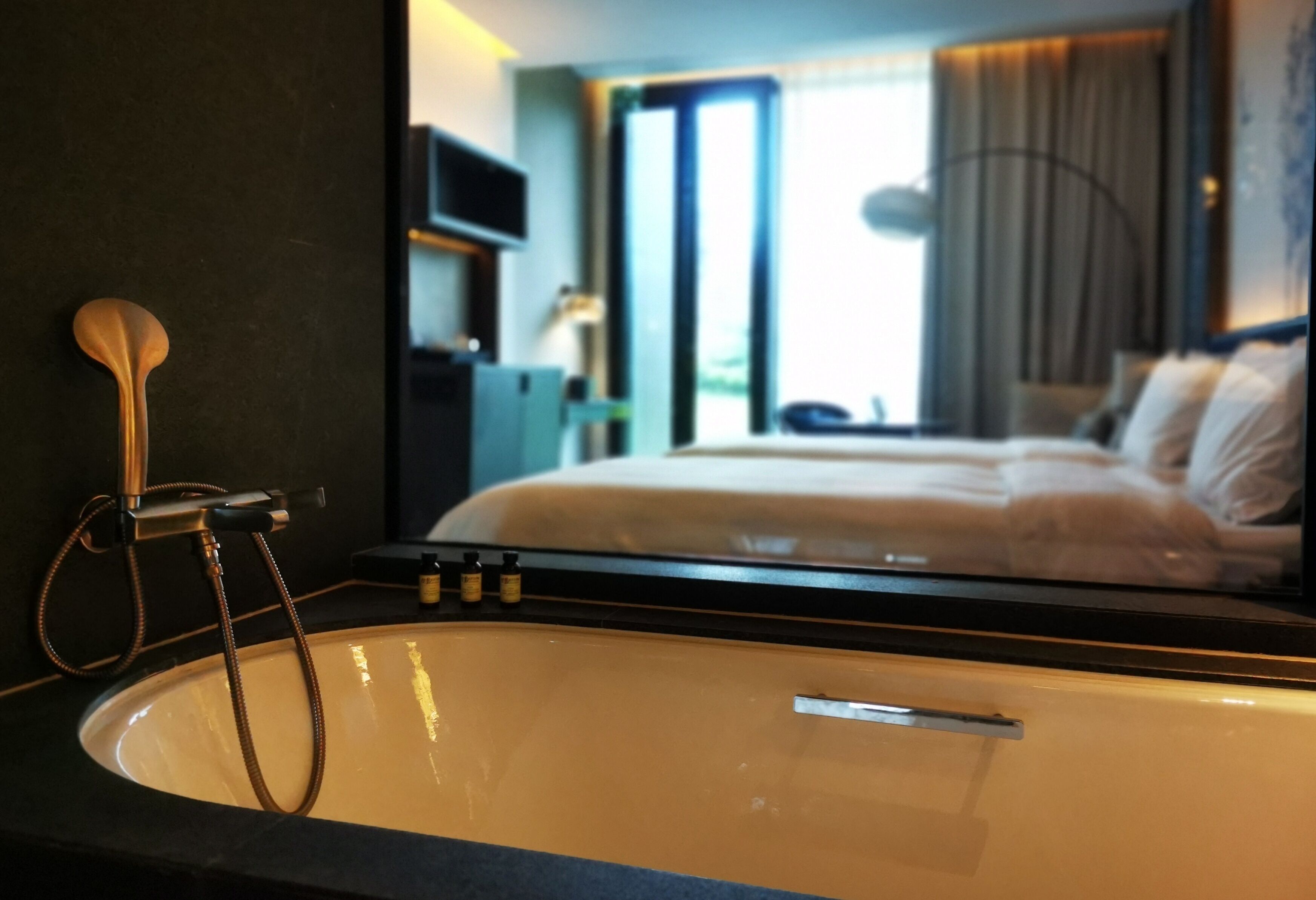Un baño con bañera, toallero y un espejo que refleja un dormitorio con cama y cortinas.