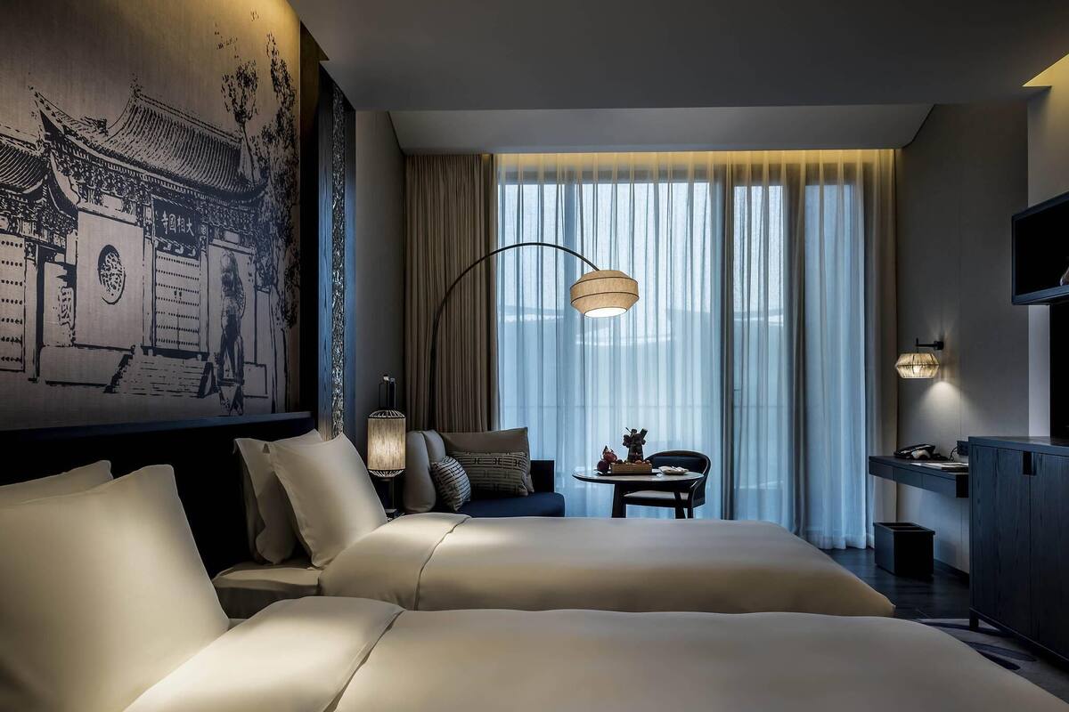 Habitación de hotel con dos camas, un escritorio, una silla, un televisor y un ventanal grande con cortinas.