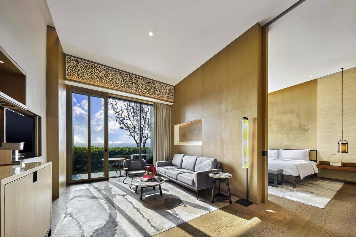 Habitación de hotel moderna con un amplio ventanal corredero que da a un balcón, un sofá, una mesa de centro, un televisor y una cama con una mesita de noche.