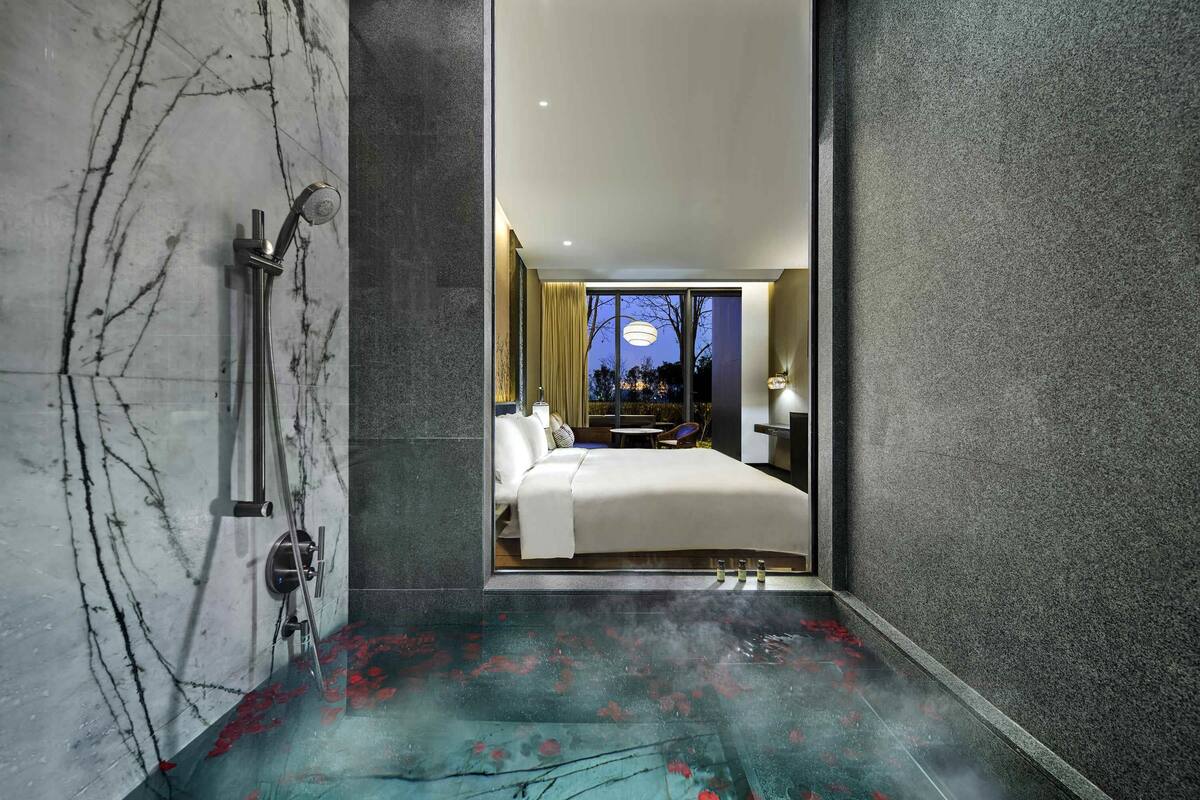 Un baño moderno con suelo de mármol, zona de ducha y un dormitorio visible con cama y mesita de noche.
