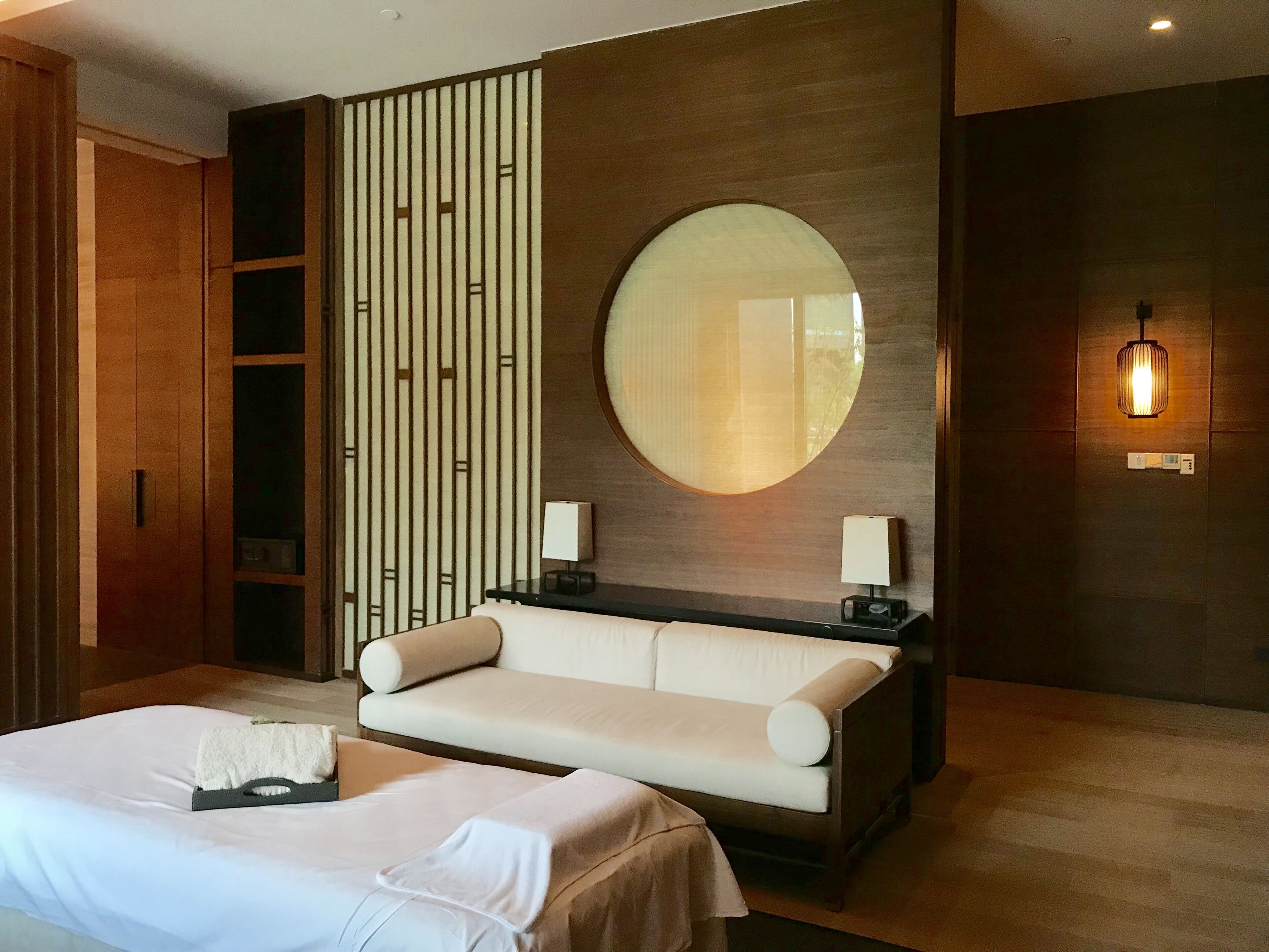 A modern hotel room with a large bed, a sofa, a round mirror, and a wall-mounted light fixture.