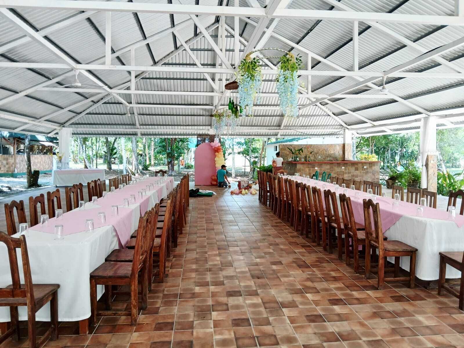 A covered dining area with long tables and wooden chairs, a tiled floor, and a high ceiling with metal beams.