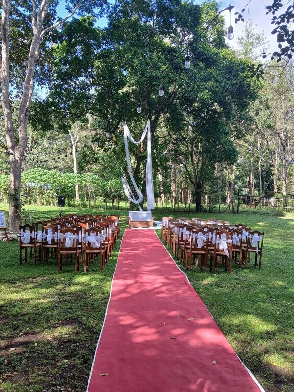 A red carpet aisle leading to a white altar with draped fabric, surrounded by wooden chairs with white cloths, set in a lush green outdoor area. - Hotel Campestre Santa Ana (Santa Ana)