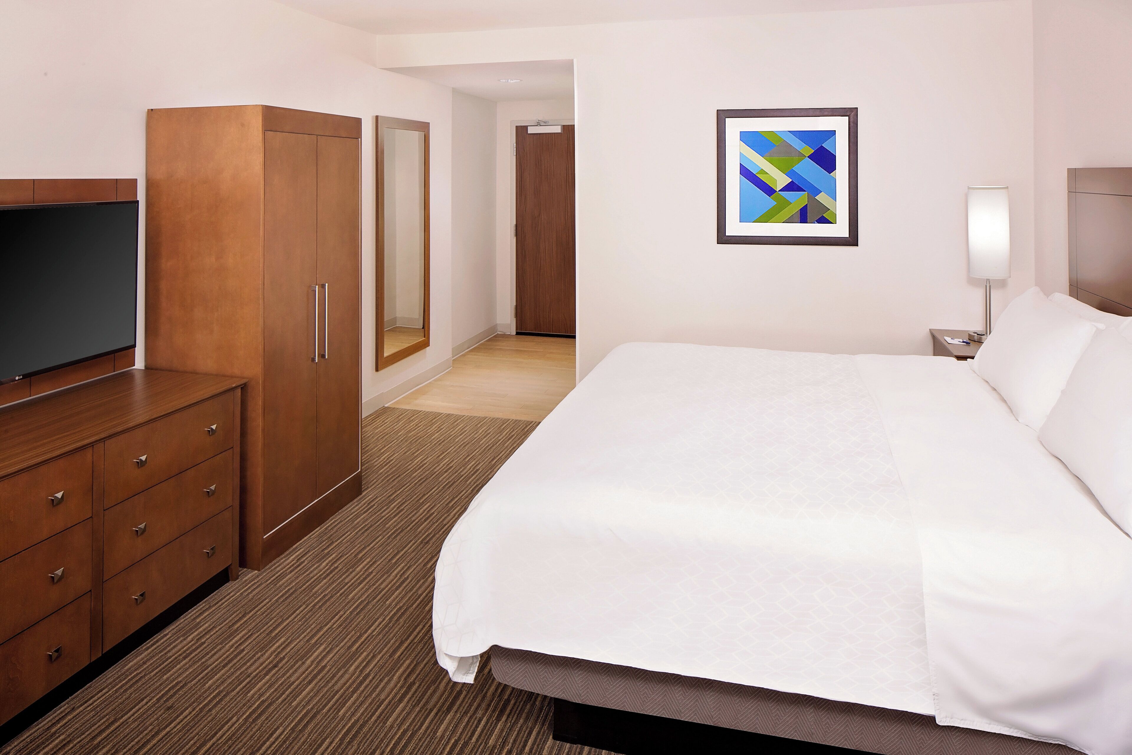 A hotel room with a bed, a TV, a wardrobe, a mirror, a framed picture, and a nightstand with a lamp.