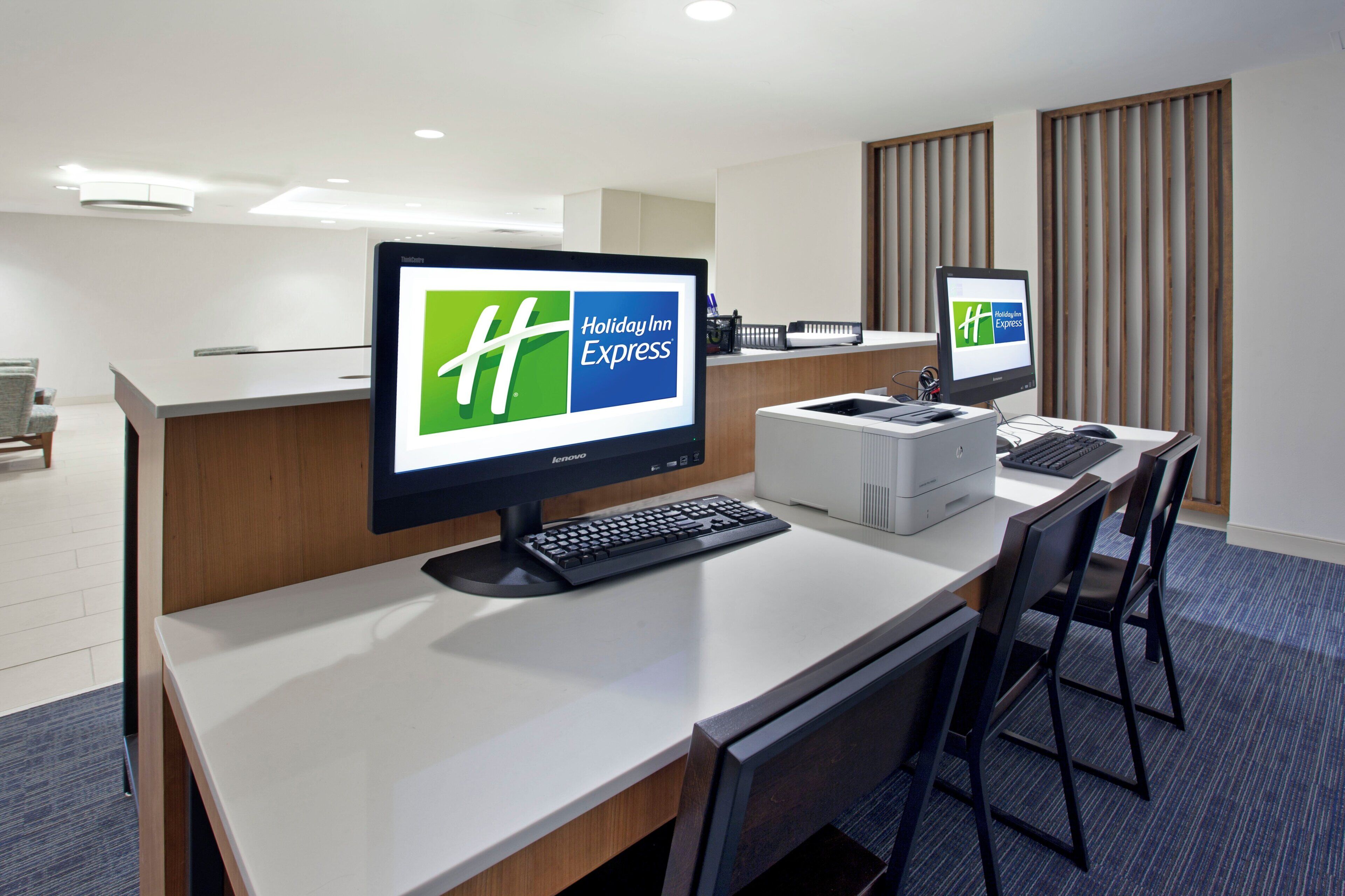 A hotel reception area with two monitors displaying the Holiday Inn Express logo, a desktop computer, a printer, and a seating area with chairs.