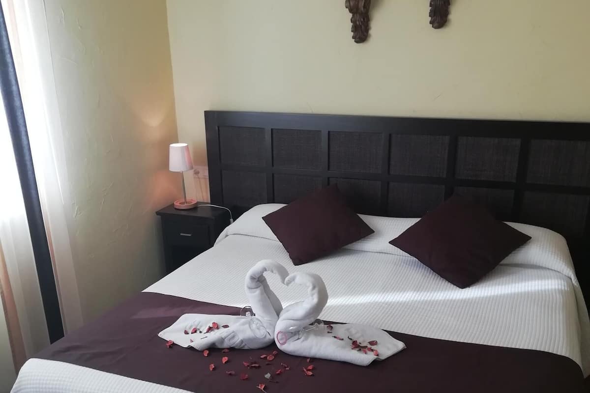 Een net opgemaakt bed met een donker hoofdbord, twee wandlampen en een decoratieve zwaan-handdoekopstelling op het bed.