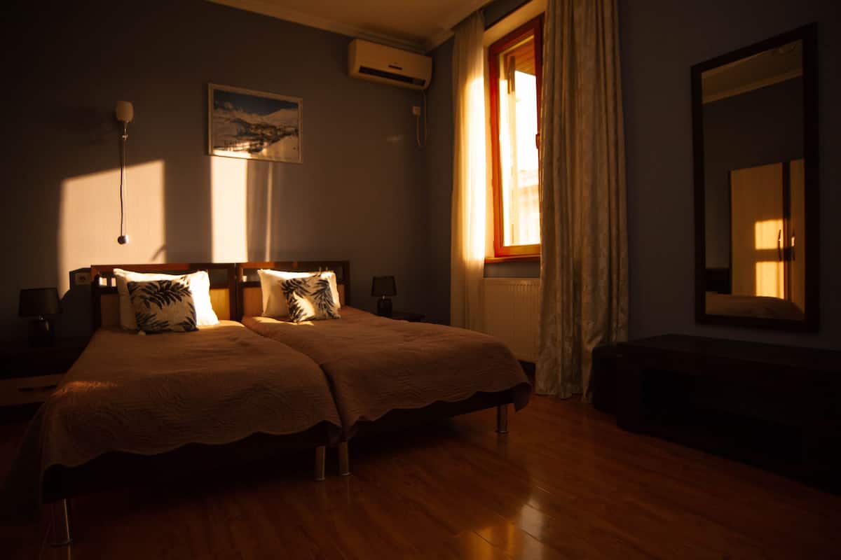 A bedroom with two beds, a nightstand, a lamp, a painting, an air conditioning unit, and a window with curtains.