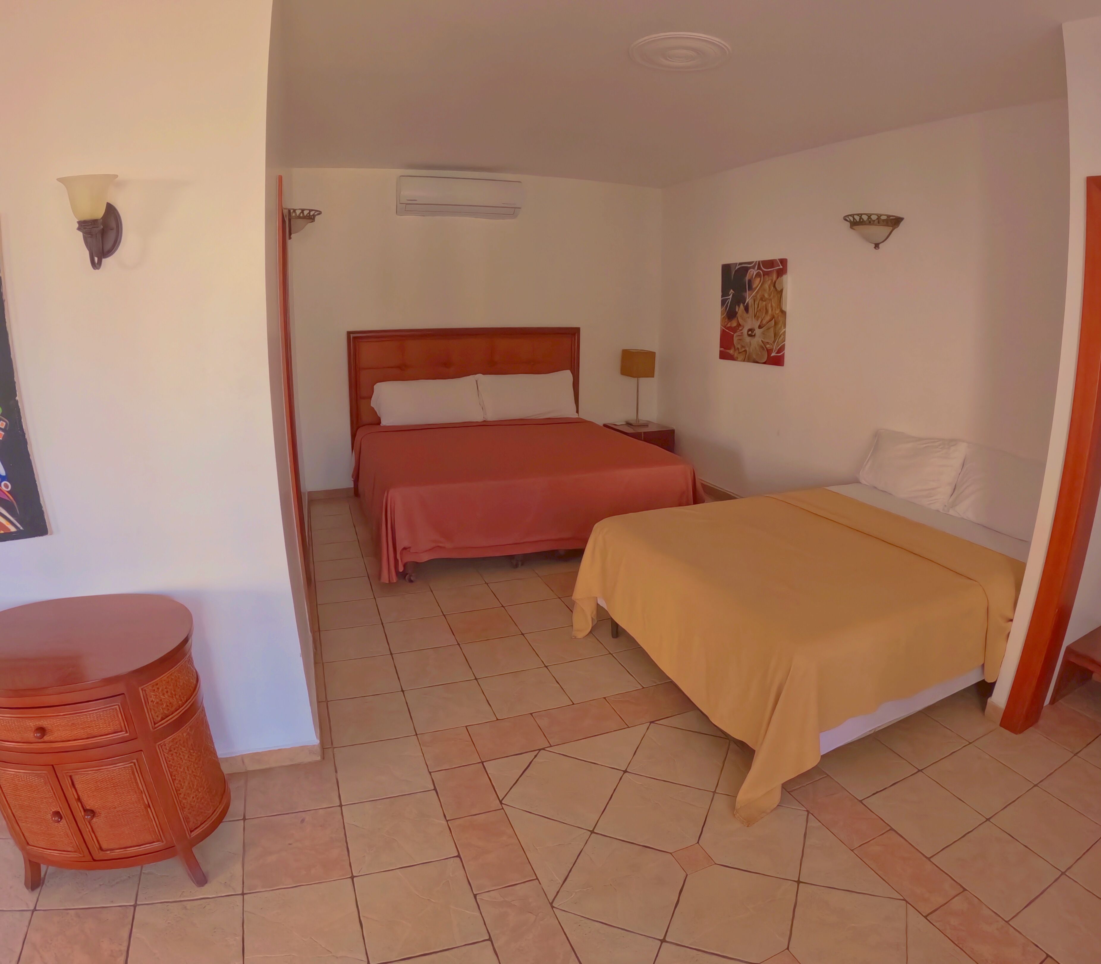 A hotel room with two beds, a wooden headboard, a small table, and a wall-mounted air conditioner.