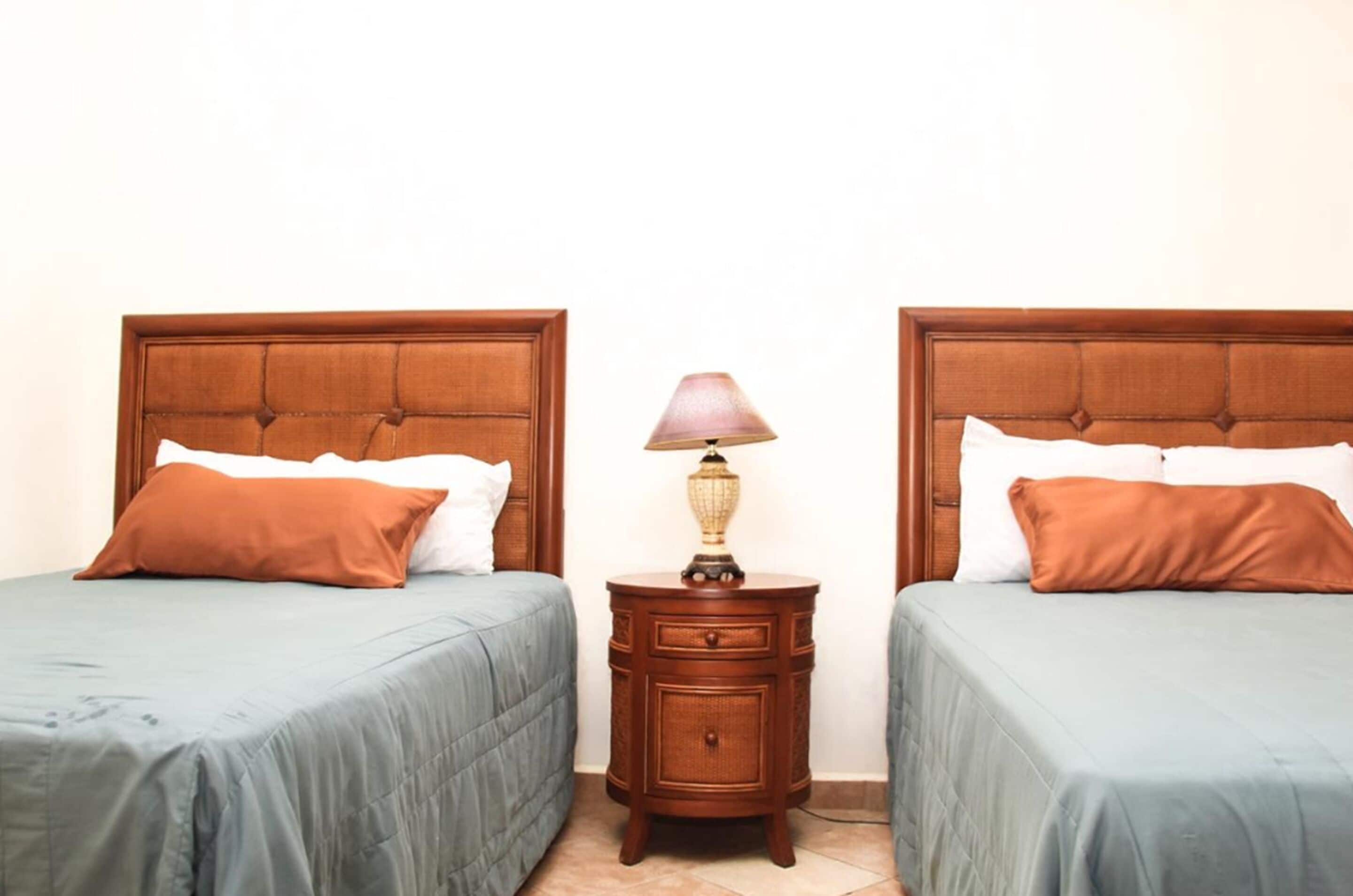 A hotel room with two beds, each with a wooden headboard, a small wooden nightstand with a lamp, and a light blue bedspread.