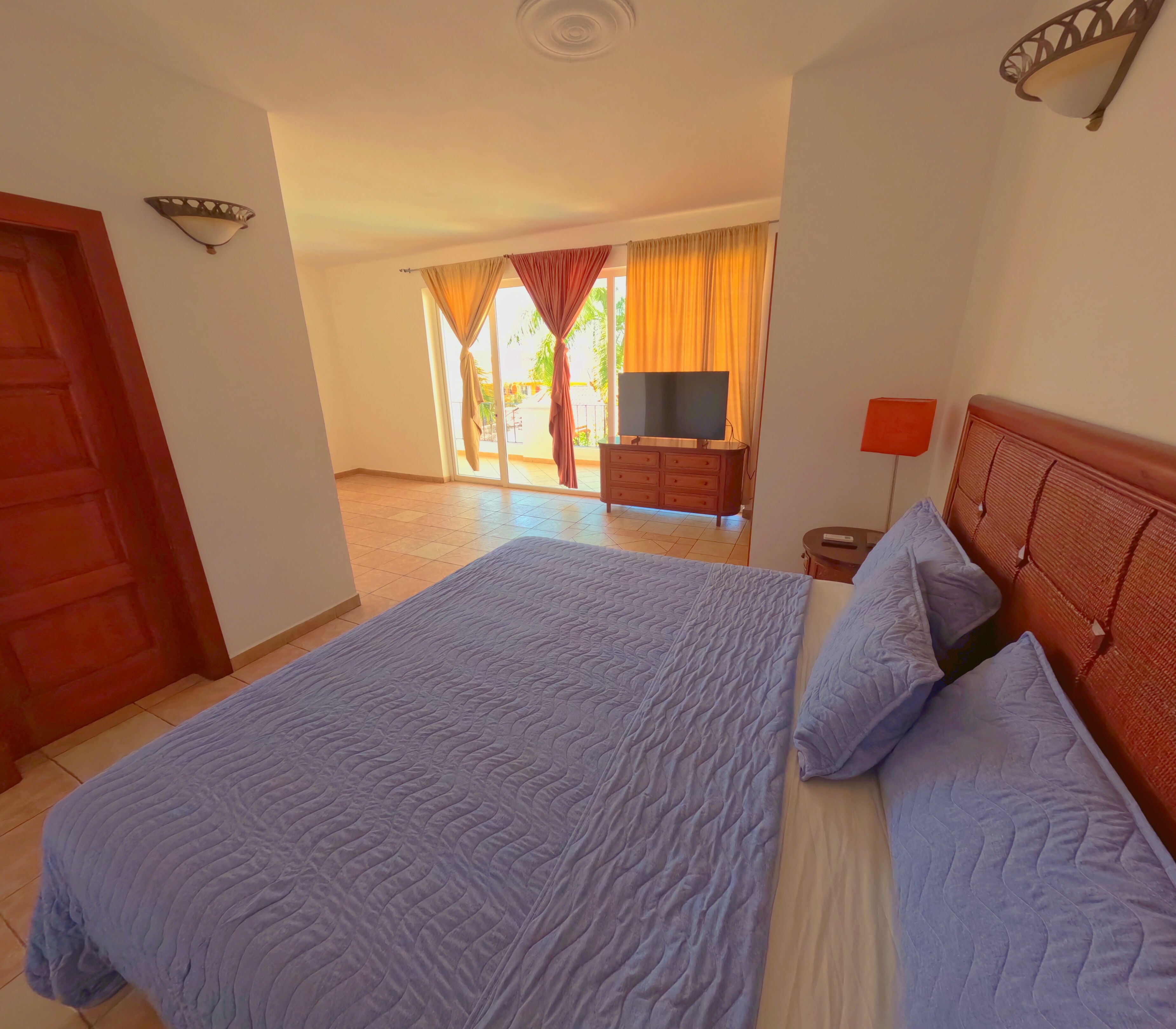 A bedroom with a large bed, a dresser, a TV, and a view of the outdoors.