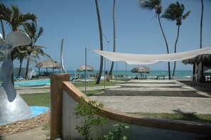 A beachside resort with a pool, lounge chairs, thatched umbrellas, and palm trees.