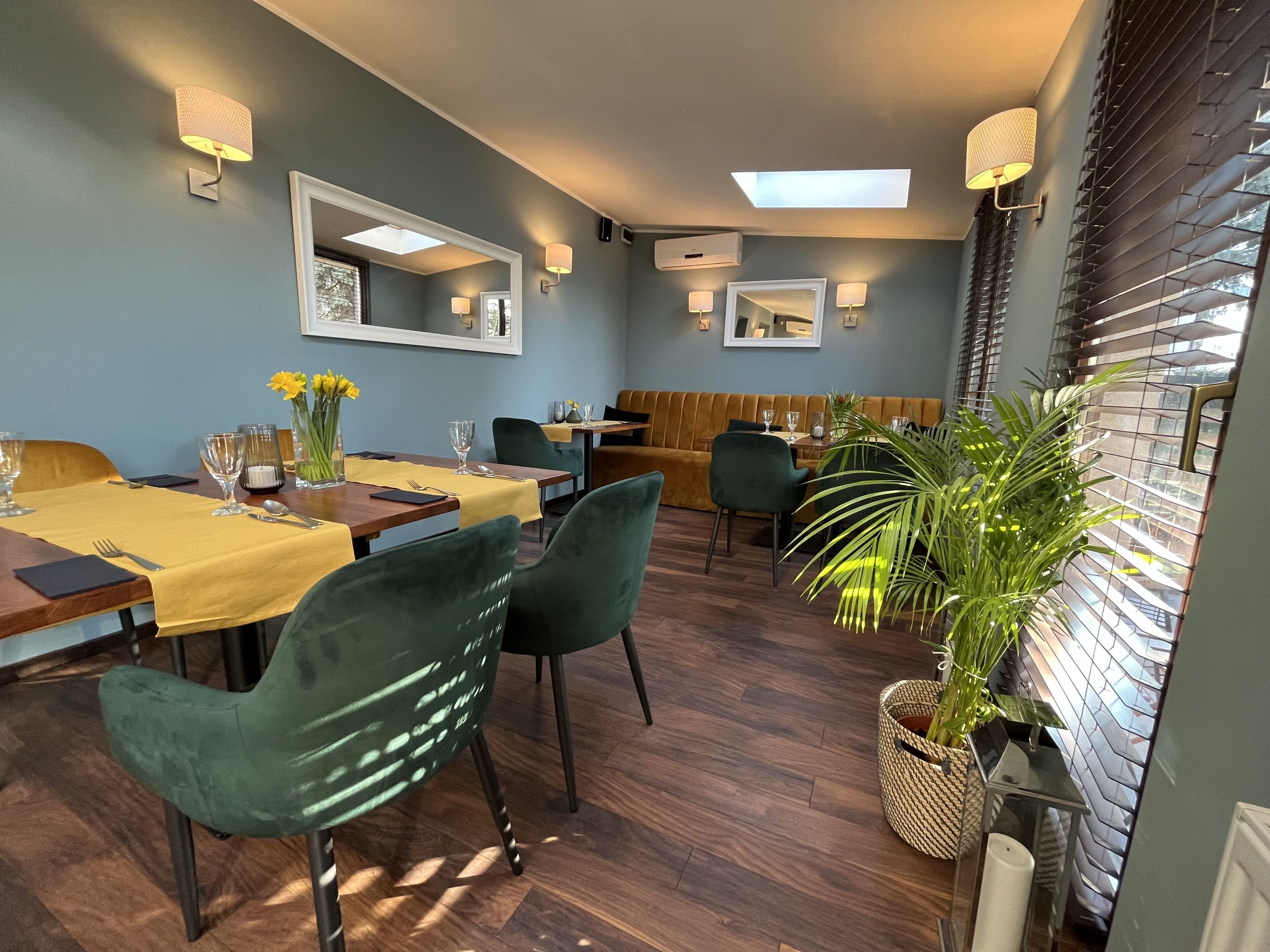 Uma área de refeições com mesas de madeira, cadeiras verdes e toalhas de mesa amarelas. Há luminárias fixadas na parede e um espelho grande.