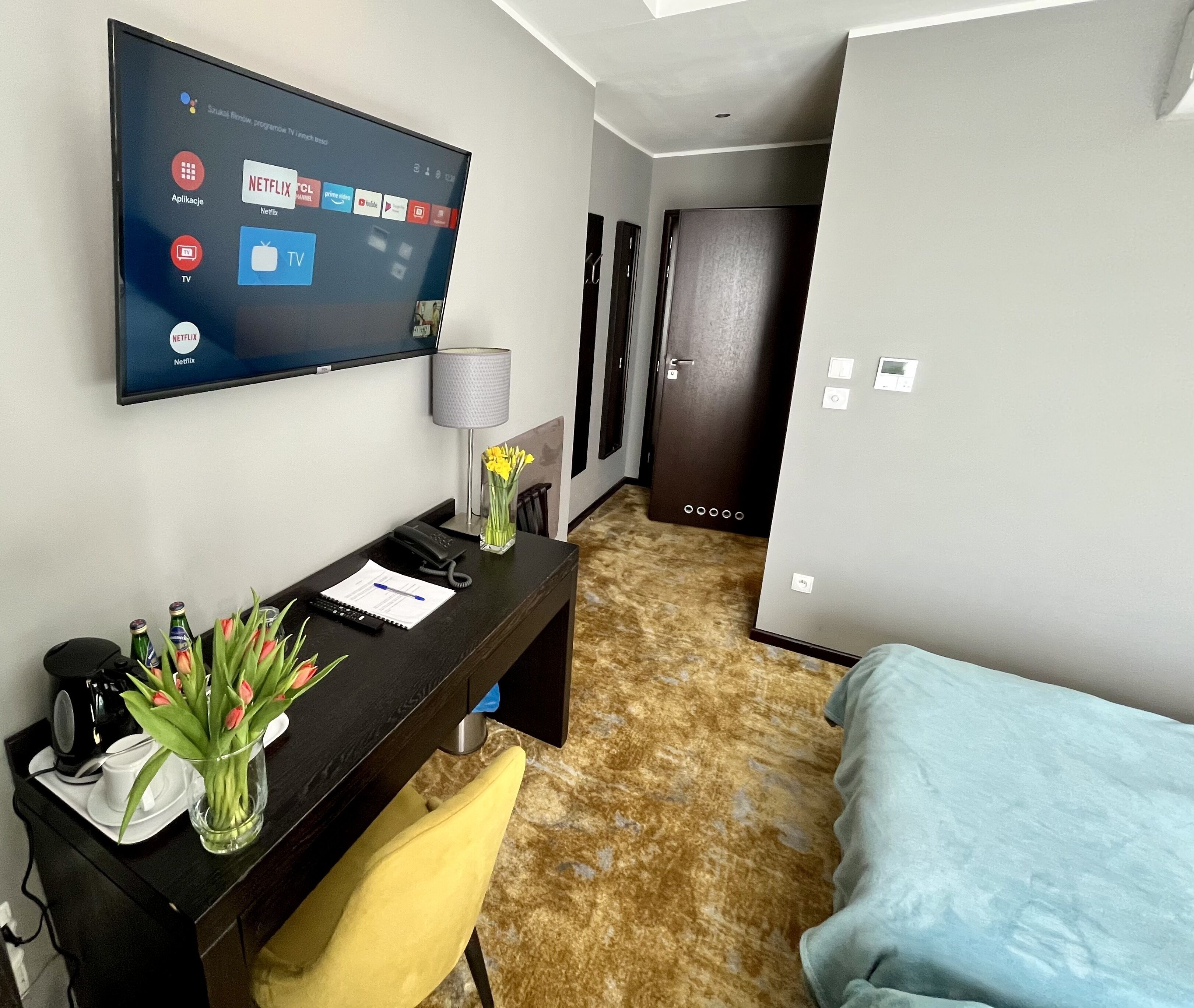 Habitación de hotel con televisión mostrando diversas aplicaciones, un escritorio con teléfono, un jarrón con flores, una silla y una cama.