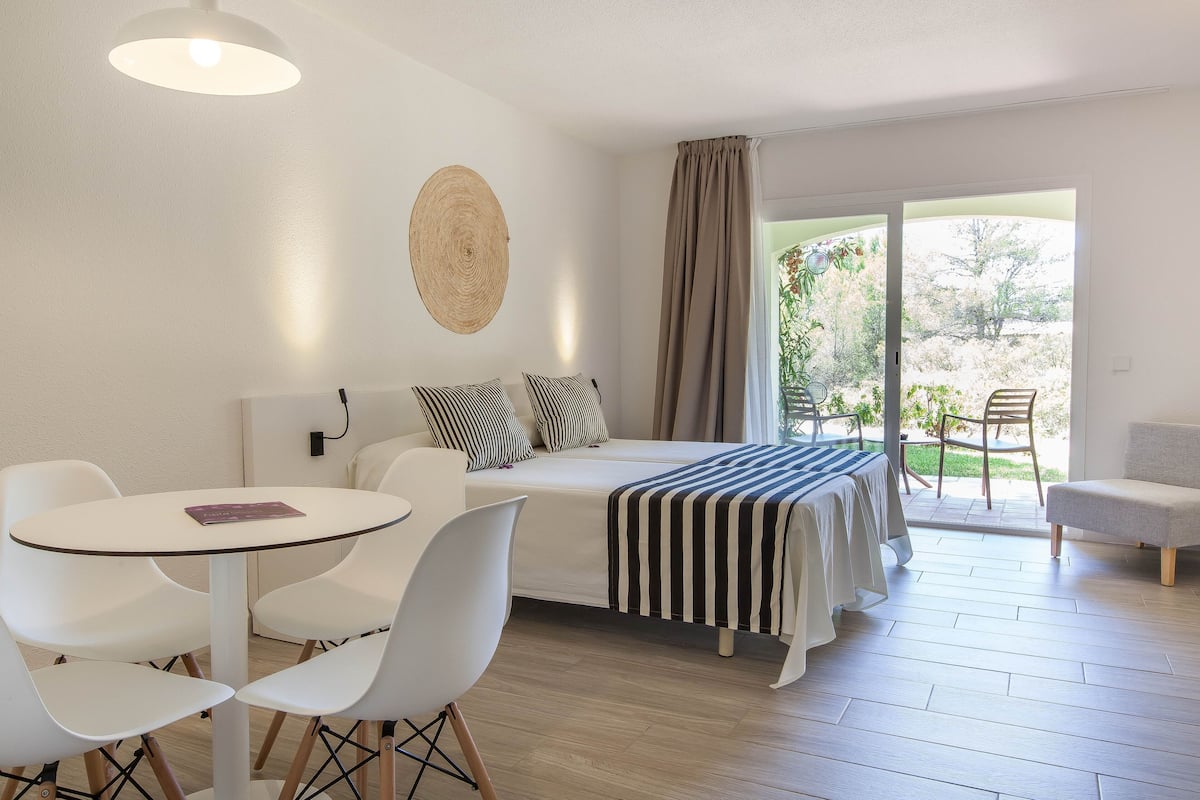 A modern room with a bed, a round wall hanging, a table with chairs, and a view of greenery outside.
