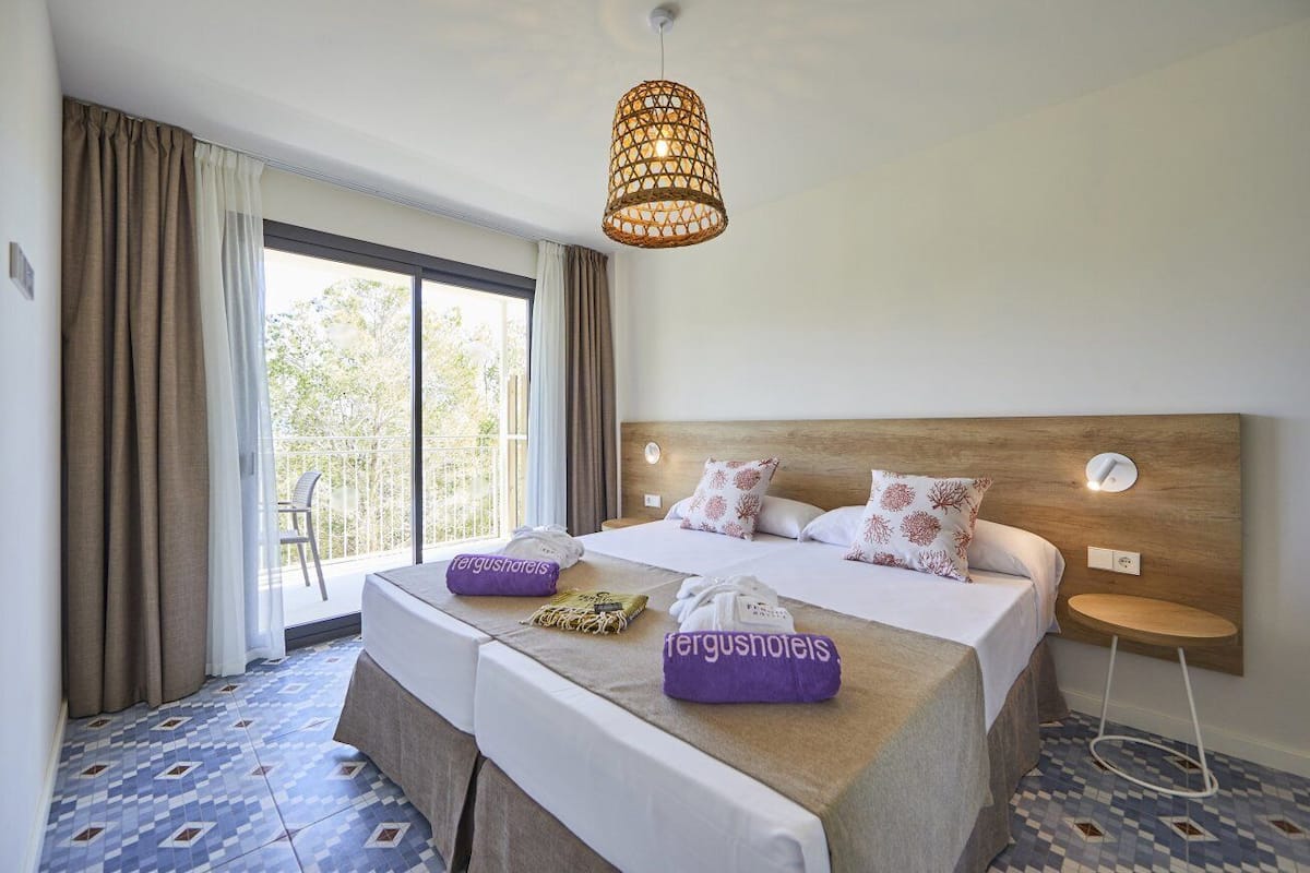 A hotel room with a large bed, a bedside table, a hanging light fixture, a window with curtains, and a view of greenery outside.