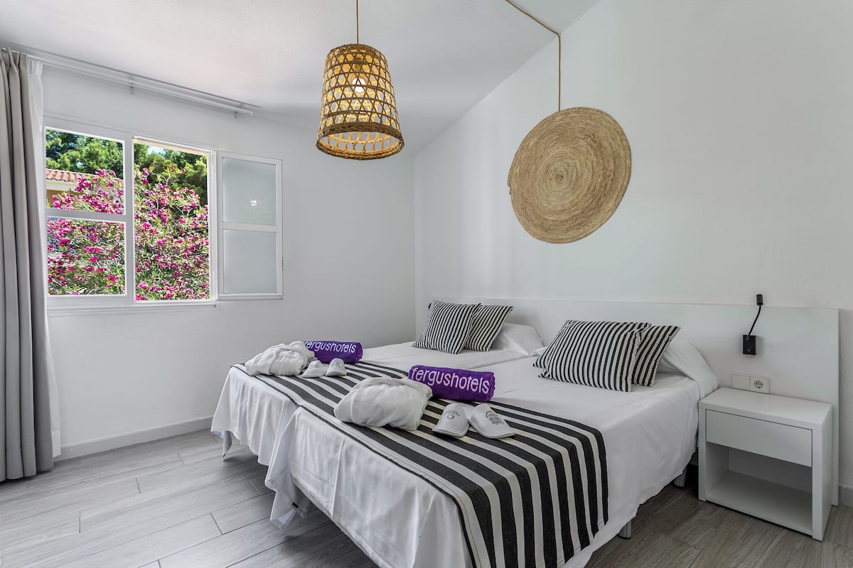 A hotel room with a bed, a bedside table, a hanging light fixture, a woven wall hanging, and a window with curtains.