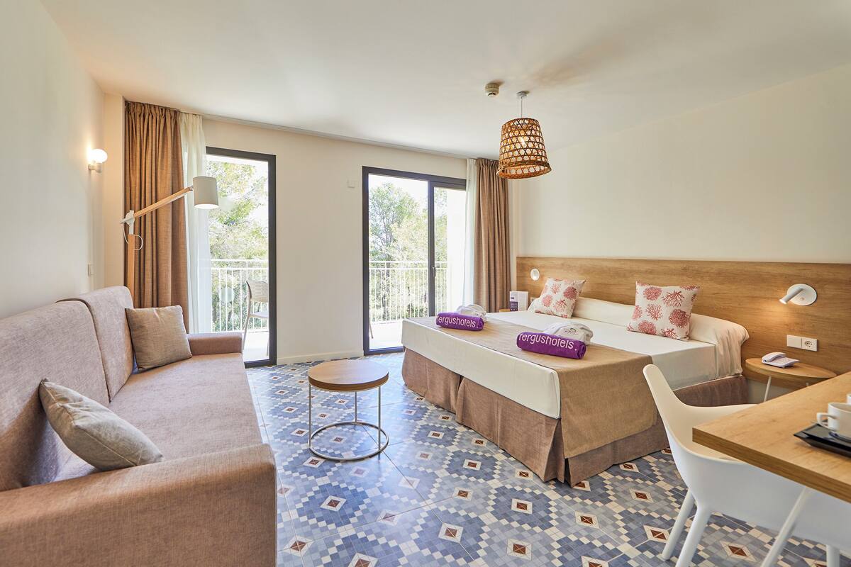 A hotel room with a sofa, a bed with a headboard, a small table, a chair, and a balcony with a view.