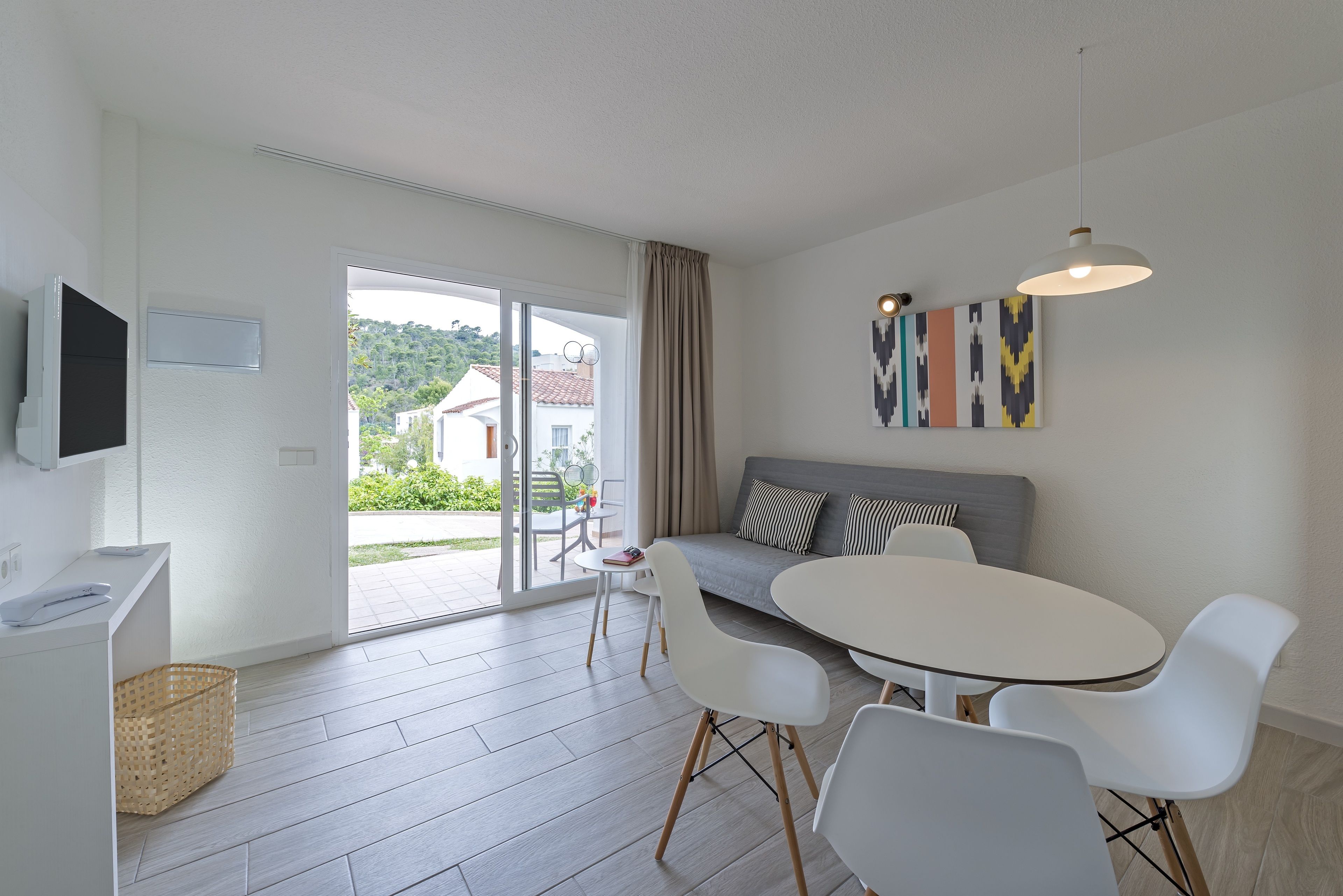 A modern living room with a sofa, a dining table, and chairs. There is a television mounted on the wall, a window with curtains, and a view of the outside.