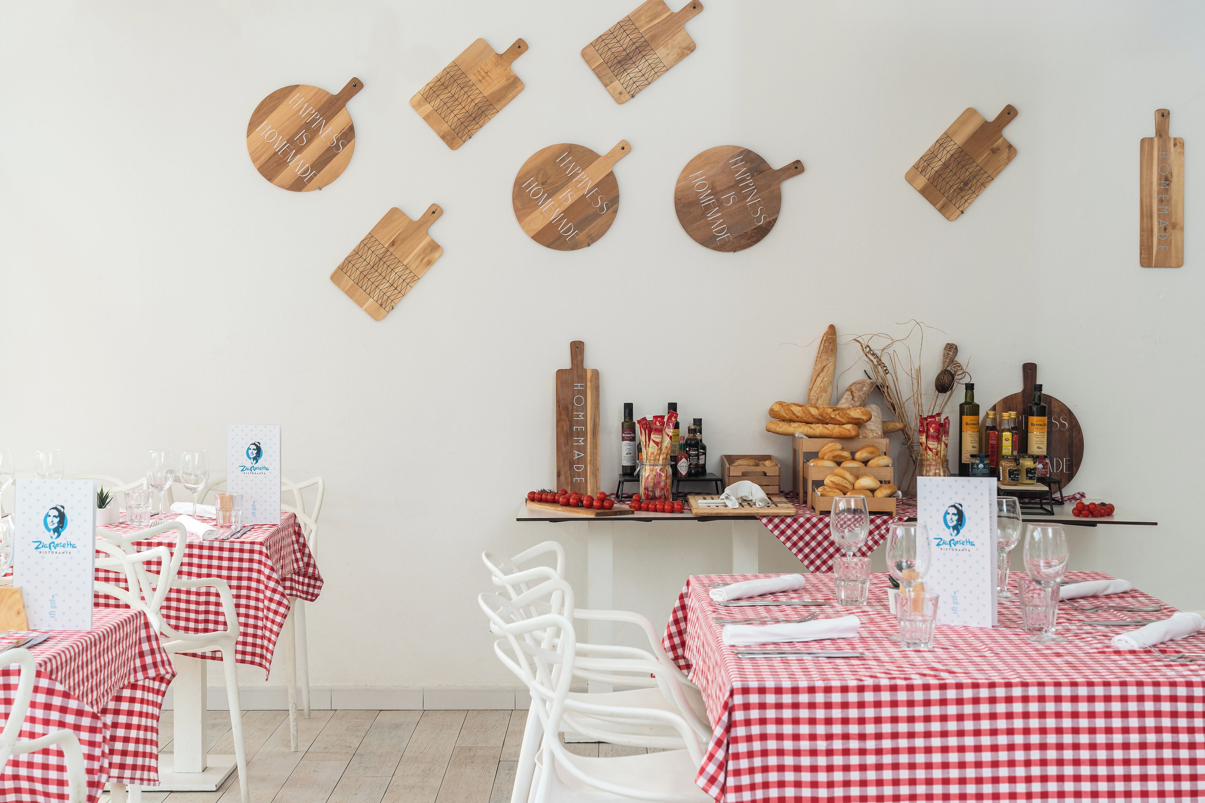 Interior de restaurante con manteles a cuadros rojos y blancos, sillas blancas y tablas de cortar de madera con texto en la pared.