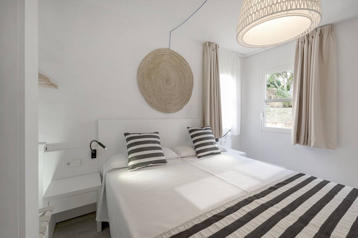 A bedroom with a large bed, two pillows, a bedside table, a lamp, a round woven wall hanging, and a window with curtains.