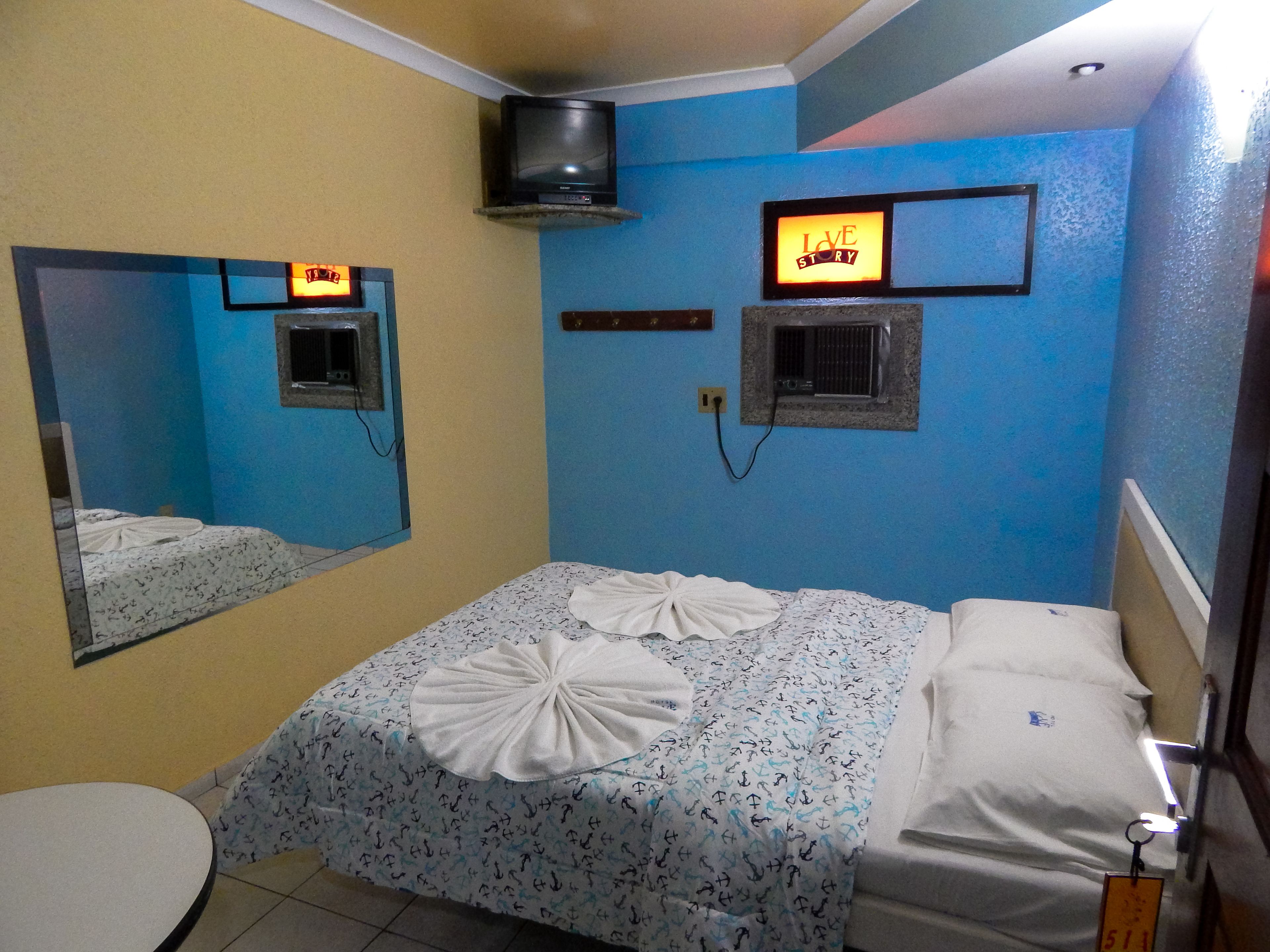 Quarto de hotel com cama, mesa de cabeceira, espelho, televisão fixada na parede e um letreiro com a inscrição 'LOVE STORY'.