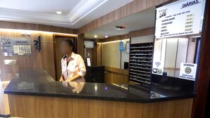 Un comptoir d’accueil d’hôtel avec un revêtement en bois et un plan de travail noir. Un panneau affiche les tarifs des chambres et les équipements disponibles, notamment le Wi-Fi, ainsi qu’un symbole de visage souriant indiquant que la caméra est en marche.