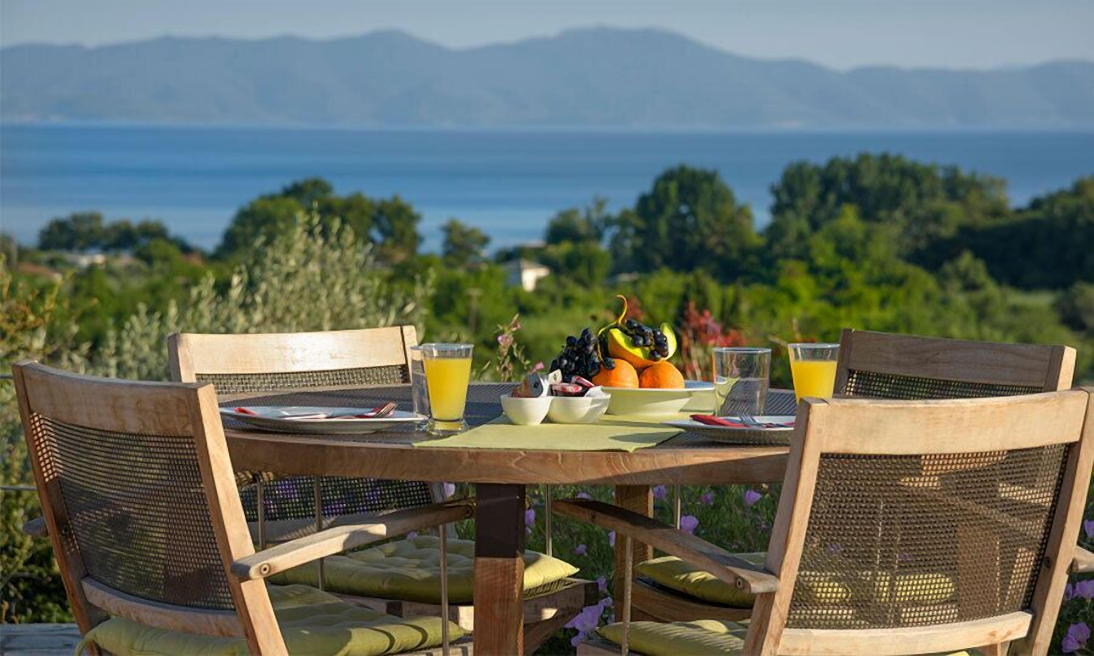 A dining table with chairs, a fruit bowl, and glasses of orange juice, set against a scenic view of a body of water and mountains.
