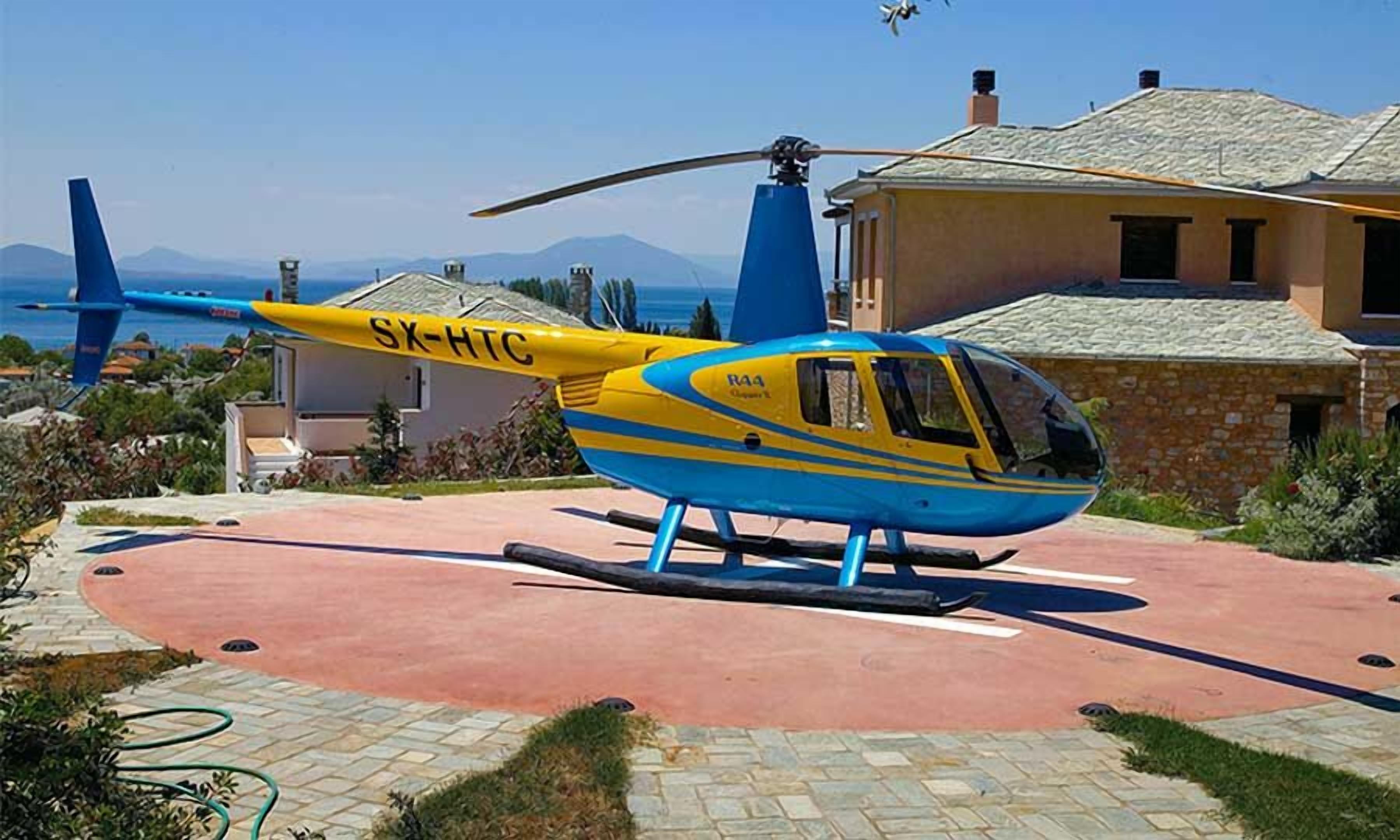 Un helicóptero azul y amarillo estacionado sobre una superficie pavimentada, con una casa y montañas al fondo.
