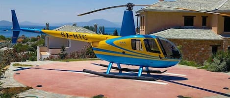 Un helicóptero azul y amarillo estacionado sobre una superficie pavimentada, con una casa y montañas al fondo.