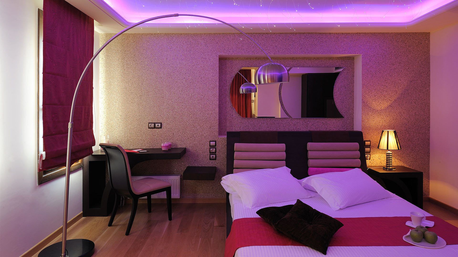 A modern hotel room with a bed, a desk, a chair, a mirror, and a floor lamp.