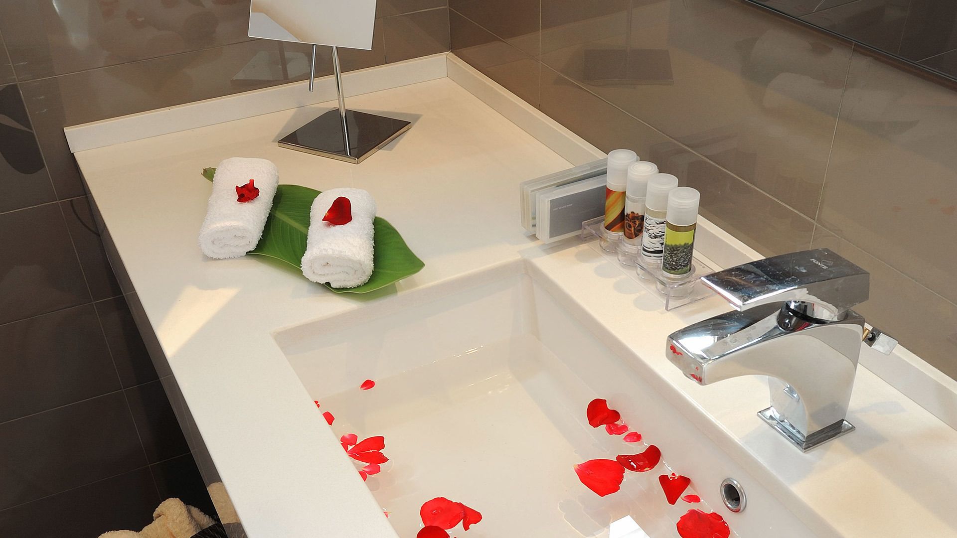 A modern bathroom with a large bathtub, two rolled towels on a green leaf, and toiletries on the side.