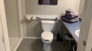 A bathroom with a toilet, a towel rack, a painting of a pier, and a stack of towels. - Smithers Guesthouse (Smithers)
