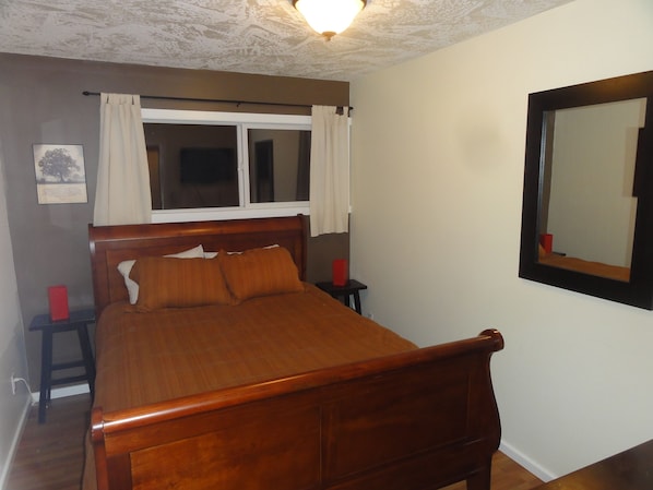 A bedroom with a wooden bed, a window with white curtains, a bedside table with a red object, and a wall-mounted mirror.
