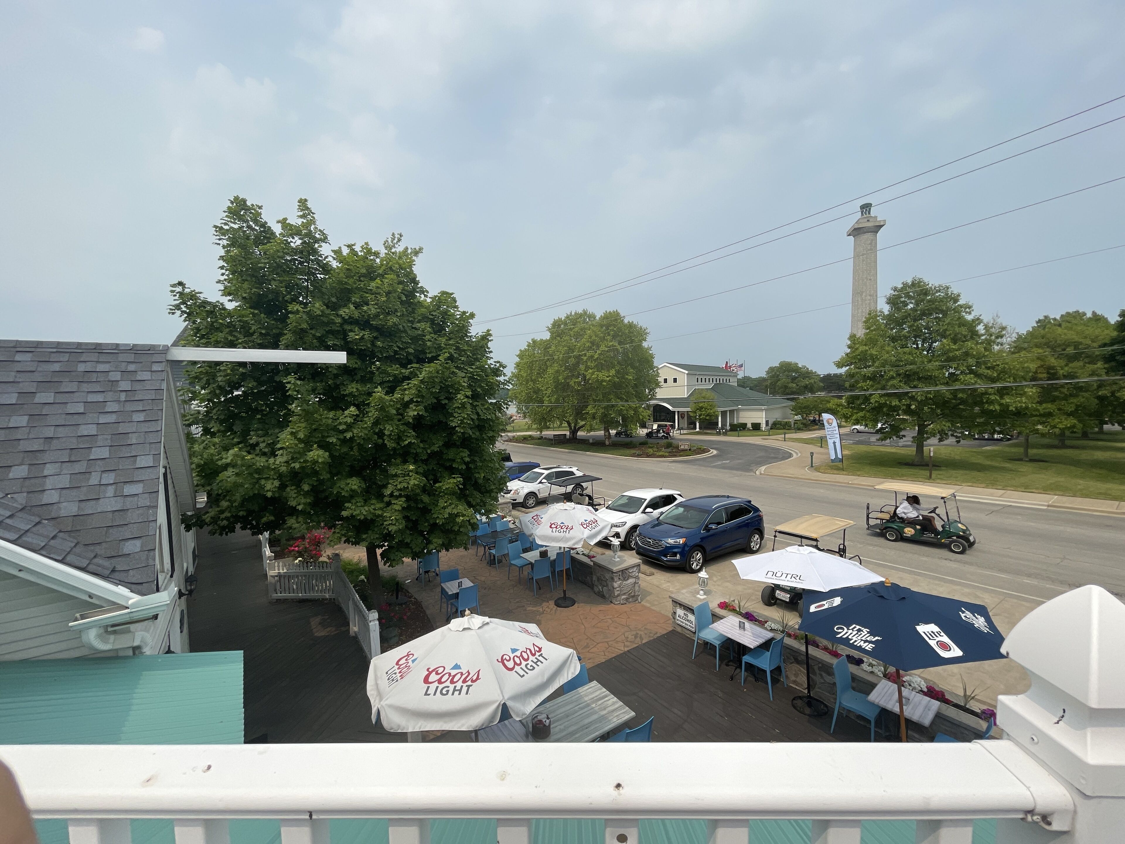 An outdoor seating area with tables and umbrellas, a parking lot with cars, and a view of a road with a flagpole and a building in the background.