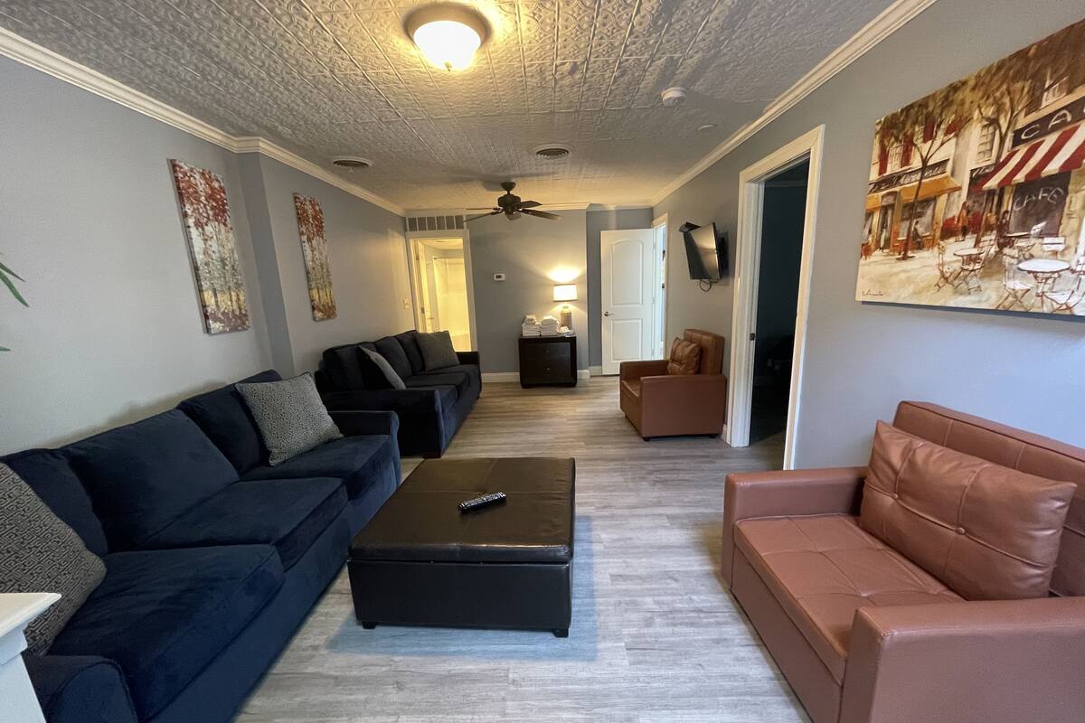 A living room with a dark blue sofa, a brown leather couch, and a black ottoman. There is a ceiling fan, a painting on the wall, and a door leading to another room.
