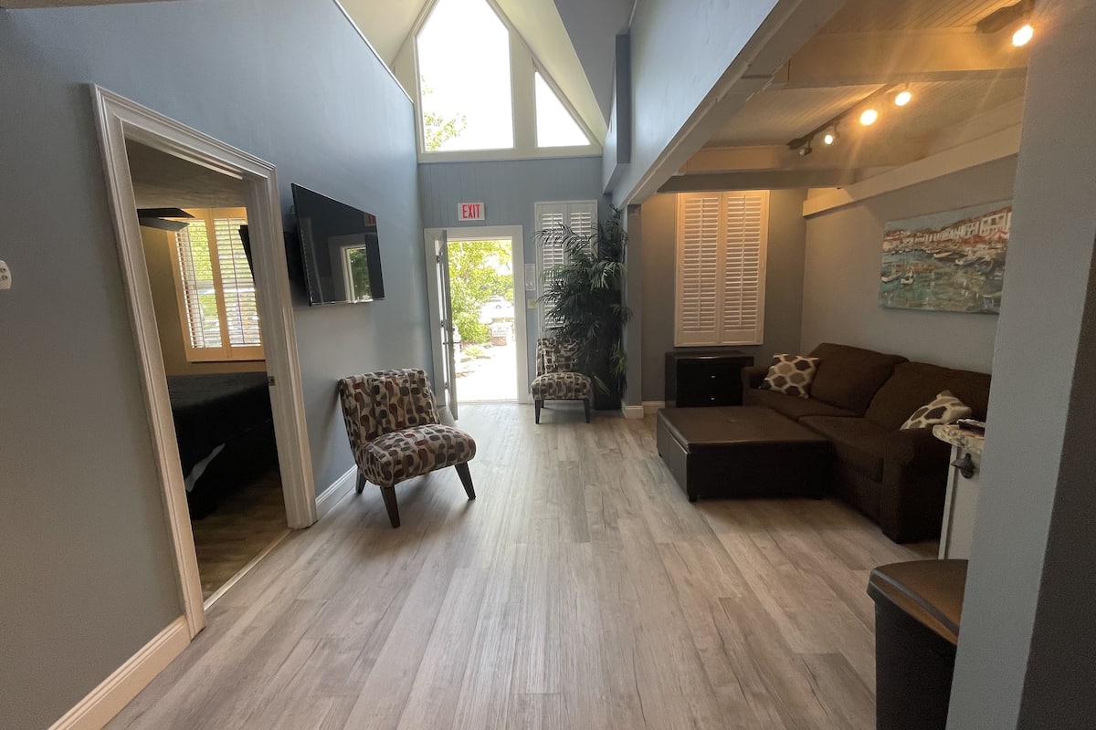 A spacious hallway with a high ceiling, a comfortable seating area with a sofa and armchairs, a television mounted on the wall, and a door leading to an outdoor area.