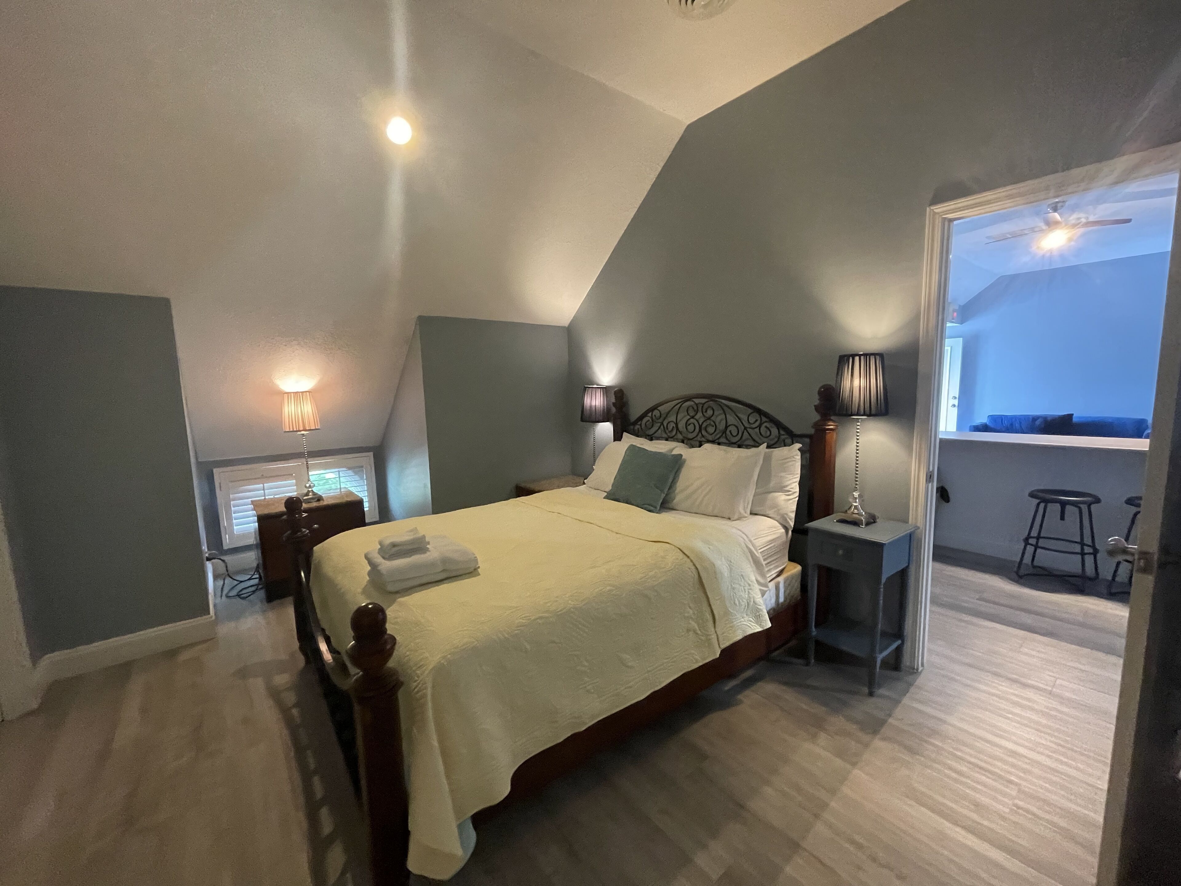 A neatly made bed with a white bedspread and pillows, a bedside table with a lamp, and a view of a balcony with a blue railing.