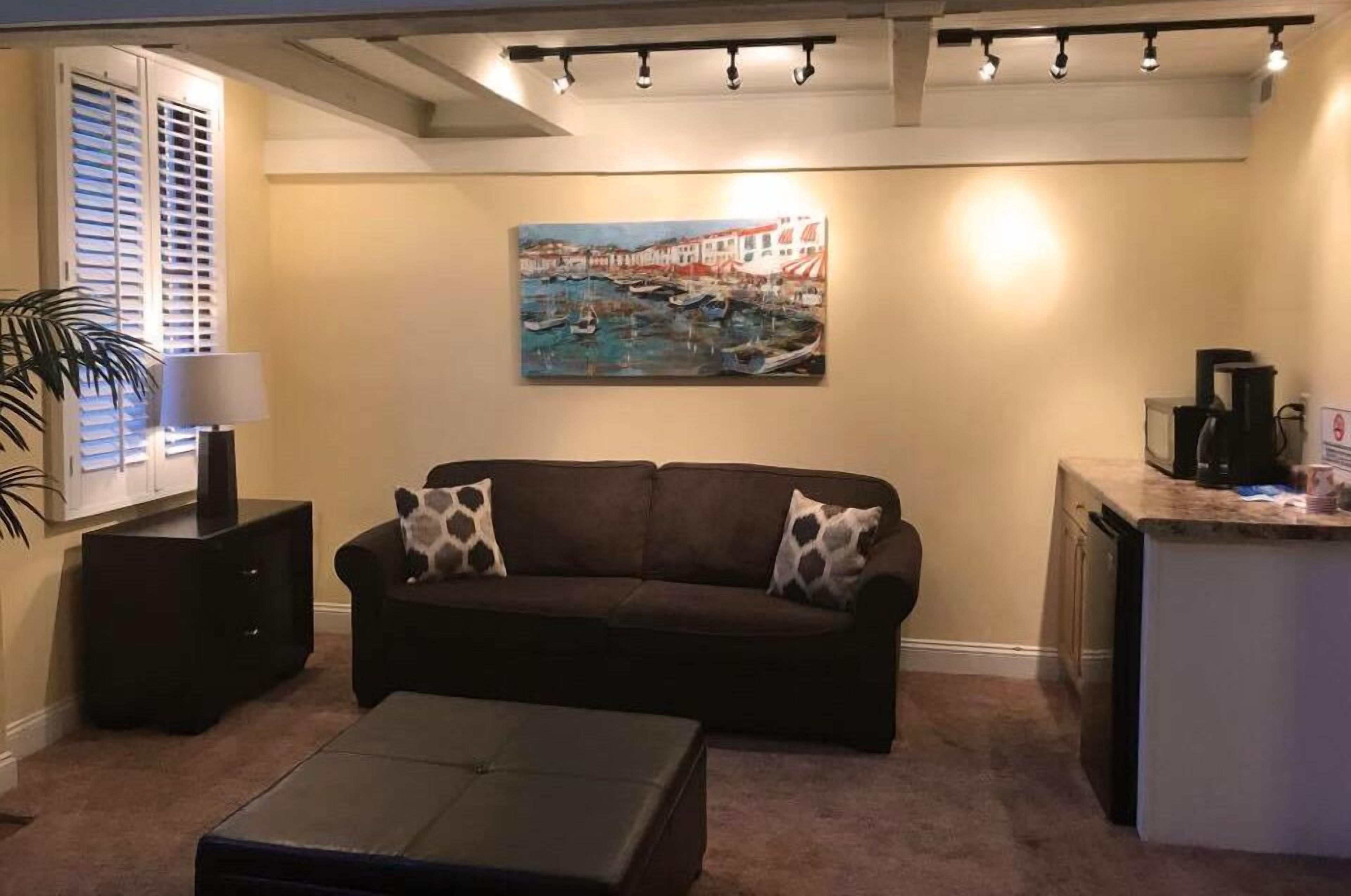 A living room with a brown sofa, two patterned pillows, a coffee table, a painting of a harbor scene, a lamp, and a small kitchen area with a microwave.