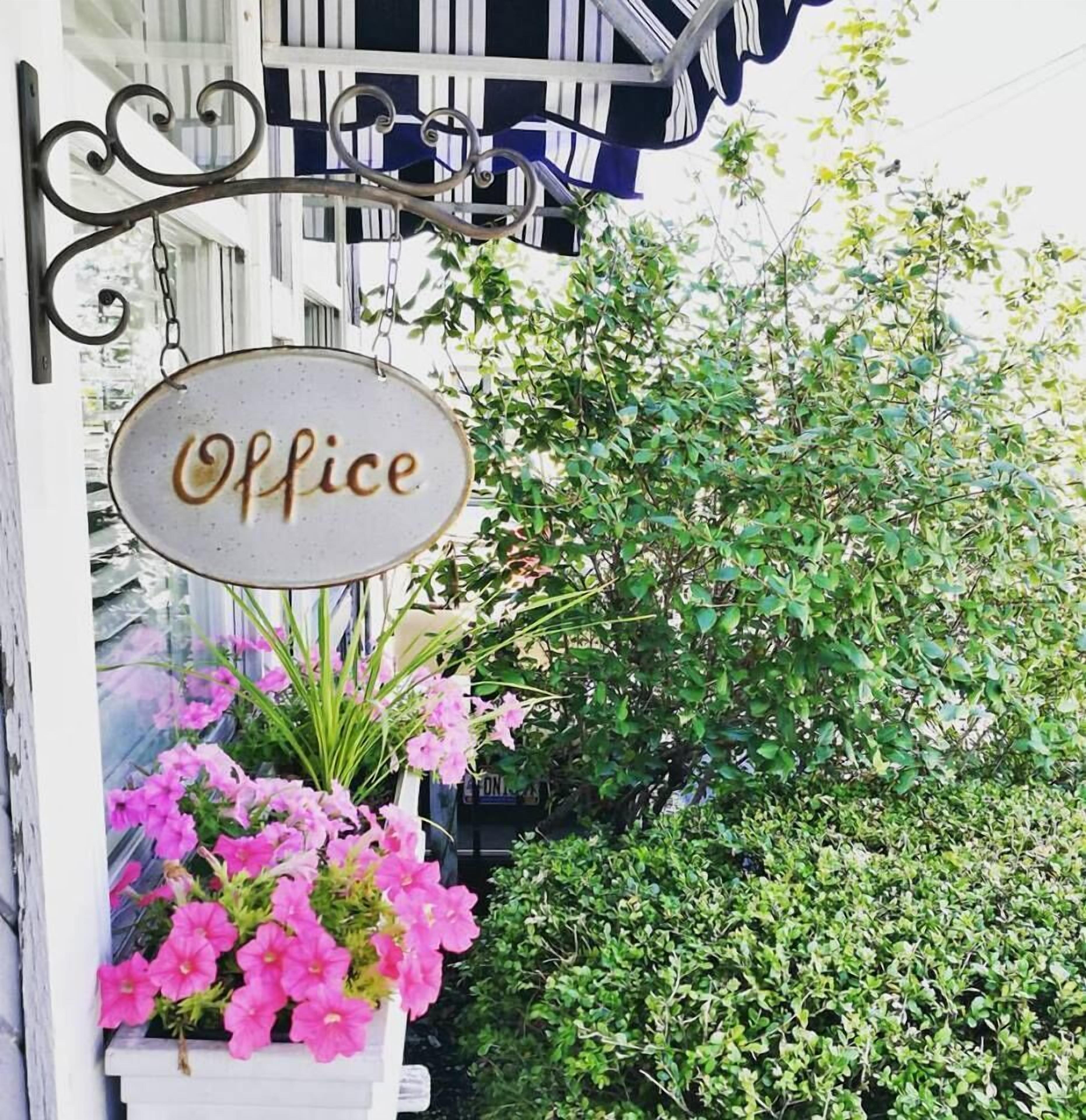 A sign reading "Office" hanging from a metal structure, with a planter of pink flowers below it.