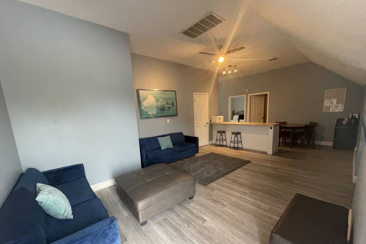 A living room with a blue sofa, a brown ottoman, and a gray rug. There is a kitchen area with a refrigerator, a microwave, and a dining table with chairs.