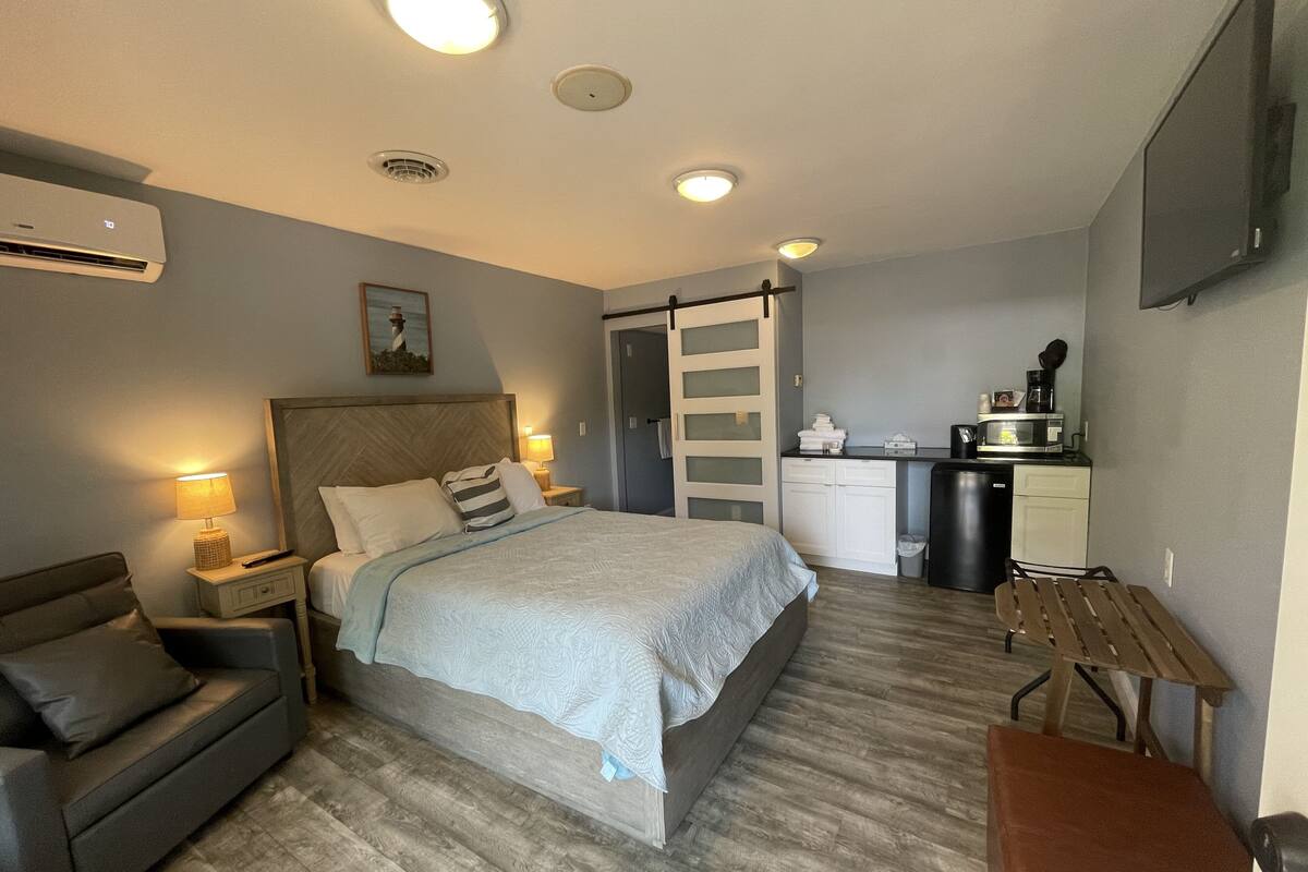A compact hotel room with a bed, bedside table, lamp, and a small seating area.