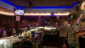 Zona de bar con pantalla de televisión, mostrador y diferentes botellas y vasos.